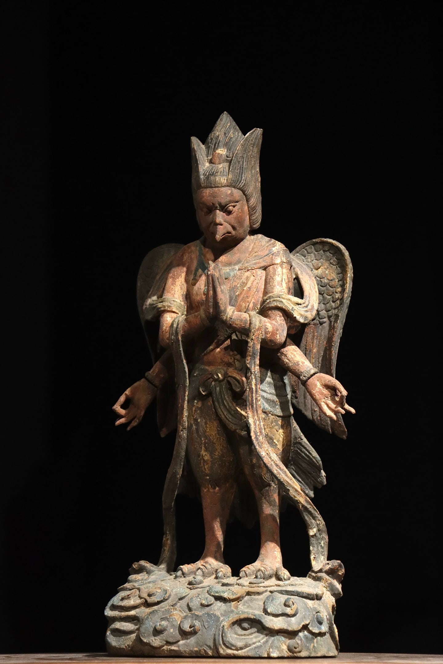 TANG DYNASTY WOODCARVING PAINTED MULTI-ARMED GARUDA: H:53cm(20.87in)Carved wood with original painted pigments. The figure has an eagle beak, human face and a pair of wings on its back, with four arms in varied gestures, standing on a cloud-patterned ba