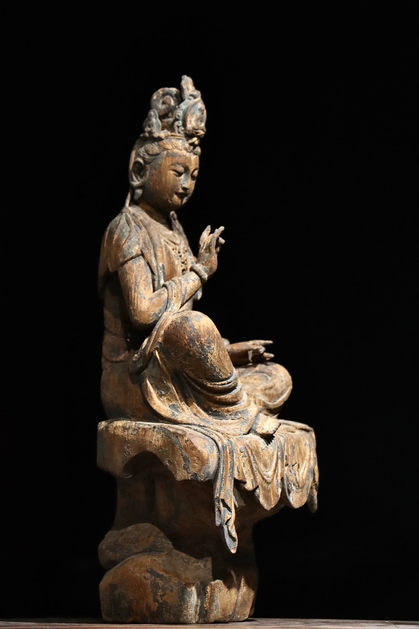 TANG DYNASTY WOODCARVING OF THE FREE AND EASY GUANYIN: H:48cm(18.90in)Carved wooden figure with an ornate crown, gentle and tranquil facial features, wearing elaborate necklaces and robes. It sits casually on a rock pedestal with natural layered drapery. 