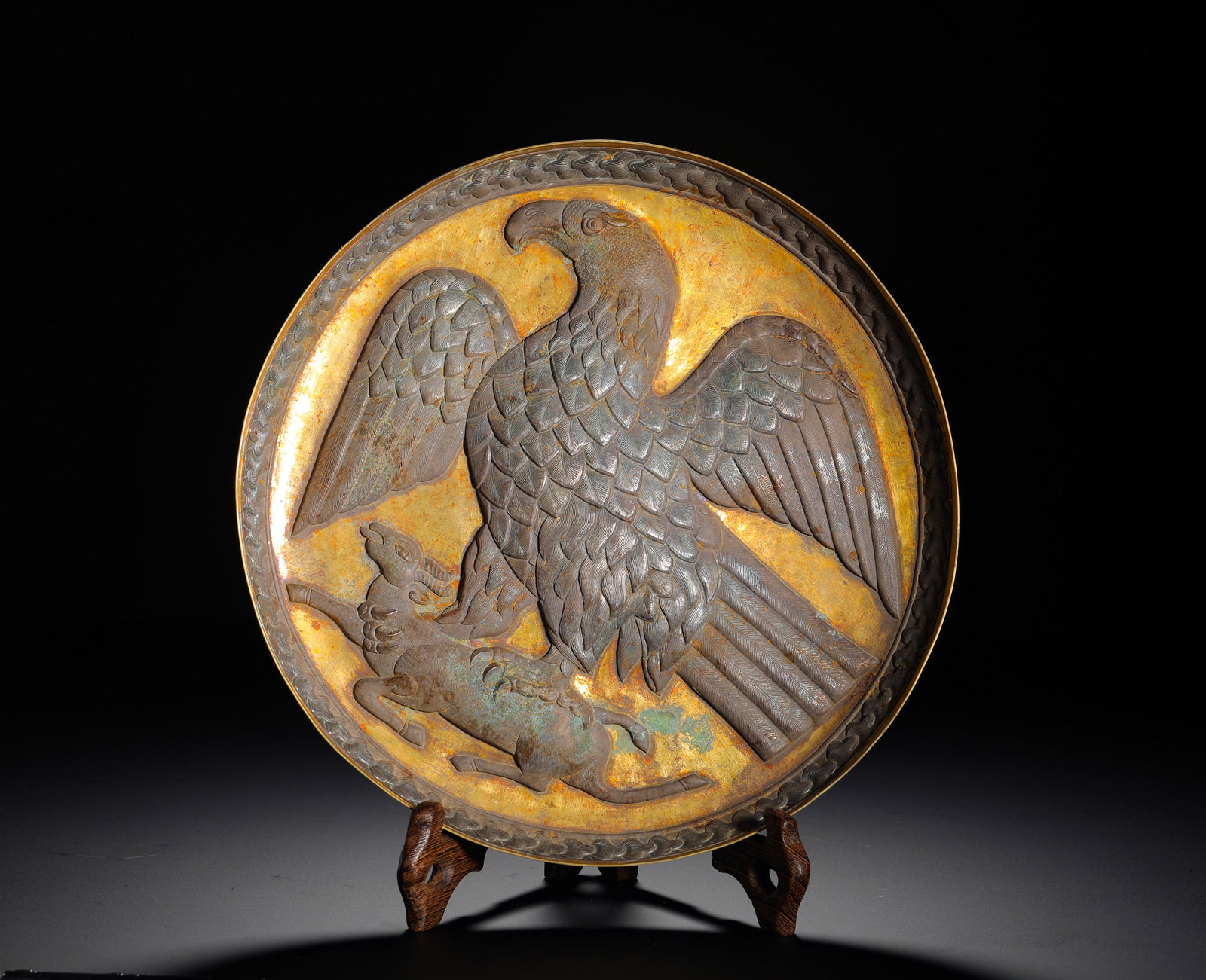 A GILDED SILVER PLATE WITH EMBOSSED EAGLE HUNTING SHEEP PATTERN FROM THE TANG DYNASTY: Height:2.5cm(0.98in) Width:22cm(8.66in) Weight:310g(0.68lb)Antique gilded silver plate embossed with an eagle capturing a sheep, interlocking vine patterns carved on rim, an ornamental display plate