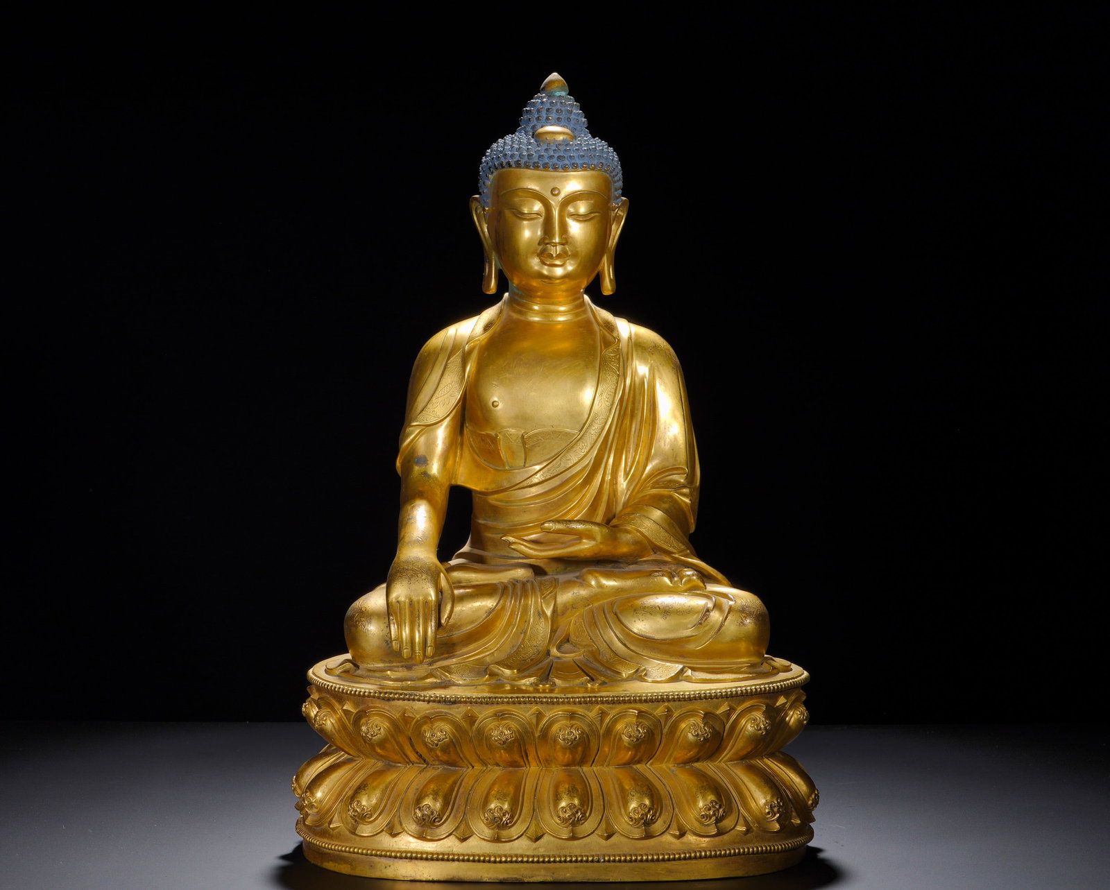 A GILDED BRONZE STATUE OF SAKYAMUNI BUDDHA ATTAINING ENLIGHTENMENT FROM THE MING DYNASTY: Height: 1cm(0.39in), Width: 33cm(12.99in), Weight: 21290g (46.936lb)His spiral ushnisha retains original blue polychrome pigment. The rounded face bears serene downcast eyes conveying calm compassion.