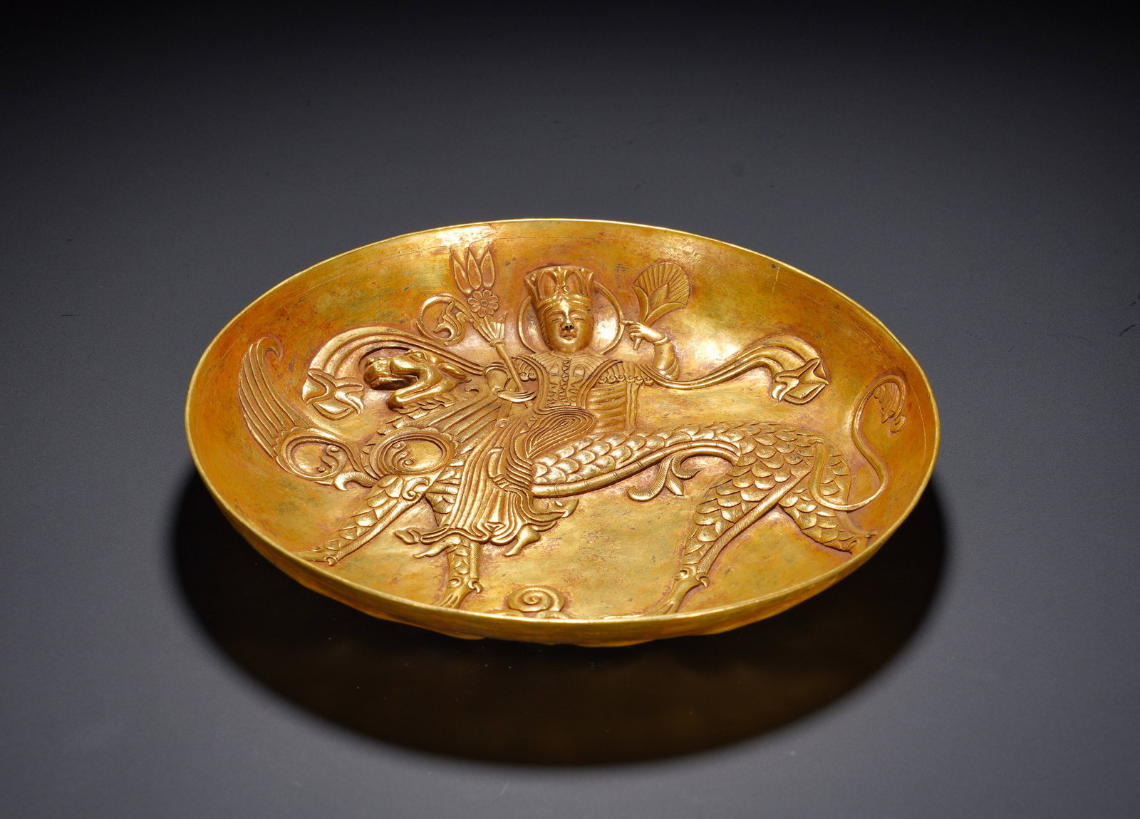 A GILDED BRONZE PLATE WITH A WINGED BEAST FIGURE PATTERN FROM THE TANG DYNASTY: Height: 3.5cm(1.38in), Diameter: 20.5cm(8.07in), Weight: 540g (1.190lb)The motif of human mounted on winged beasts derives from Silk Road cultural exchange, combining Eastern and Western iconography, 