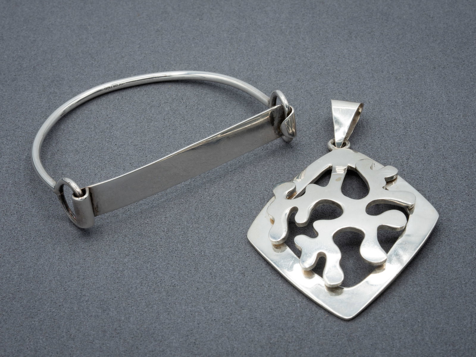 Two 925 Sterling Silver Modernist Jewelry Pieces: A two-piece grouping of Mexican modernist silver jewelry comprising an openwork pendant and a sculptural bangle style bracelet. The pendant features an abstract raised design within a rounded-square f