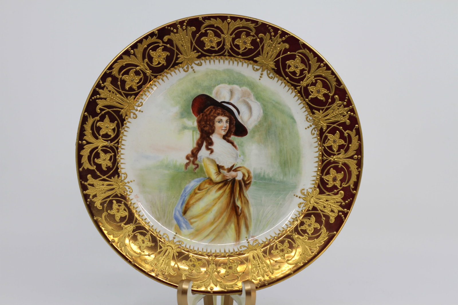 Antique Continental Hand-Painted Porcelain Portrait Cabinet Plate with Burgundy and Raised Gilt: An antique Continental European porcelain cabinet plate centered with a finely hand-painted portrait of an elegantly dressed young woman in a golden-yellow gown, white fichu and broad-brimmed feathere