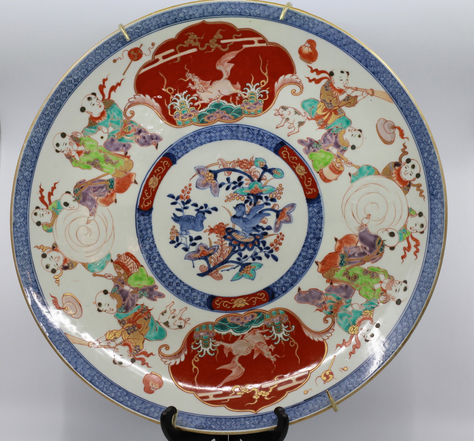 JAPANESE IMARI PORCELAIN CHARGER: Large porcelain charger decorated in underglaze blue, iron red, green, aubergine and gilt. The central reserve depicts birds among flowering branches, surrounded by playful children, cranes and auspic