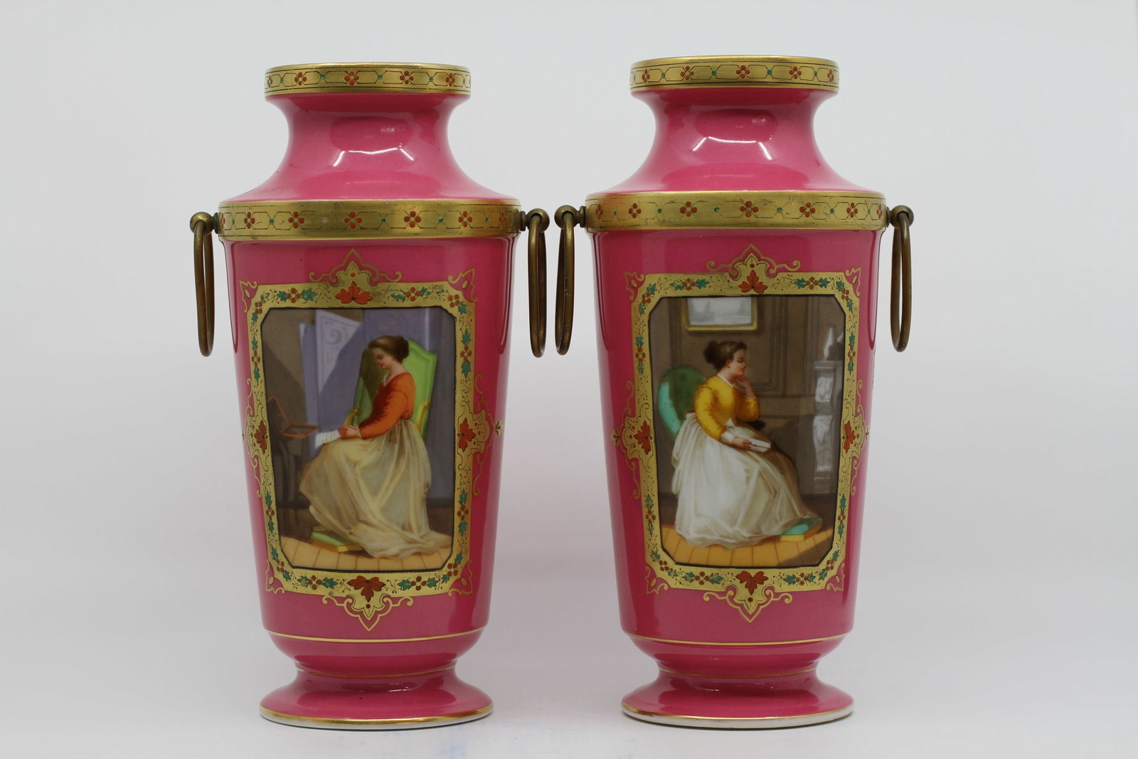 Pair of Antique Pink Porcelain Vases with Figural Scenes: Pair of antique porcelain vases with vibrant pink ground, gilt decoration, hand painted interior figural scenes, and gilt metal ring handles. Each vase features a framed portrait scene of a seated wom