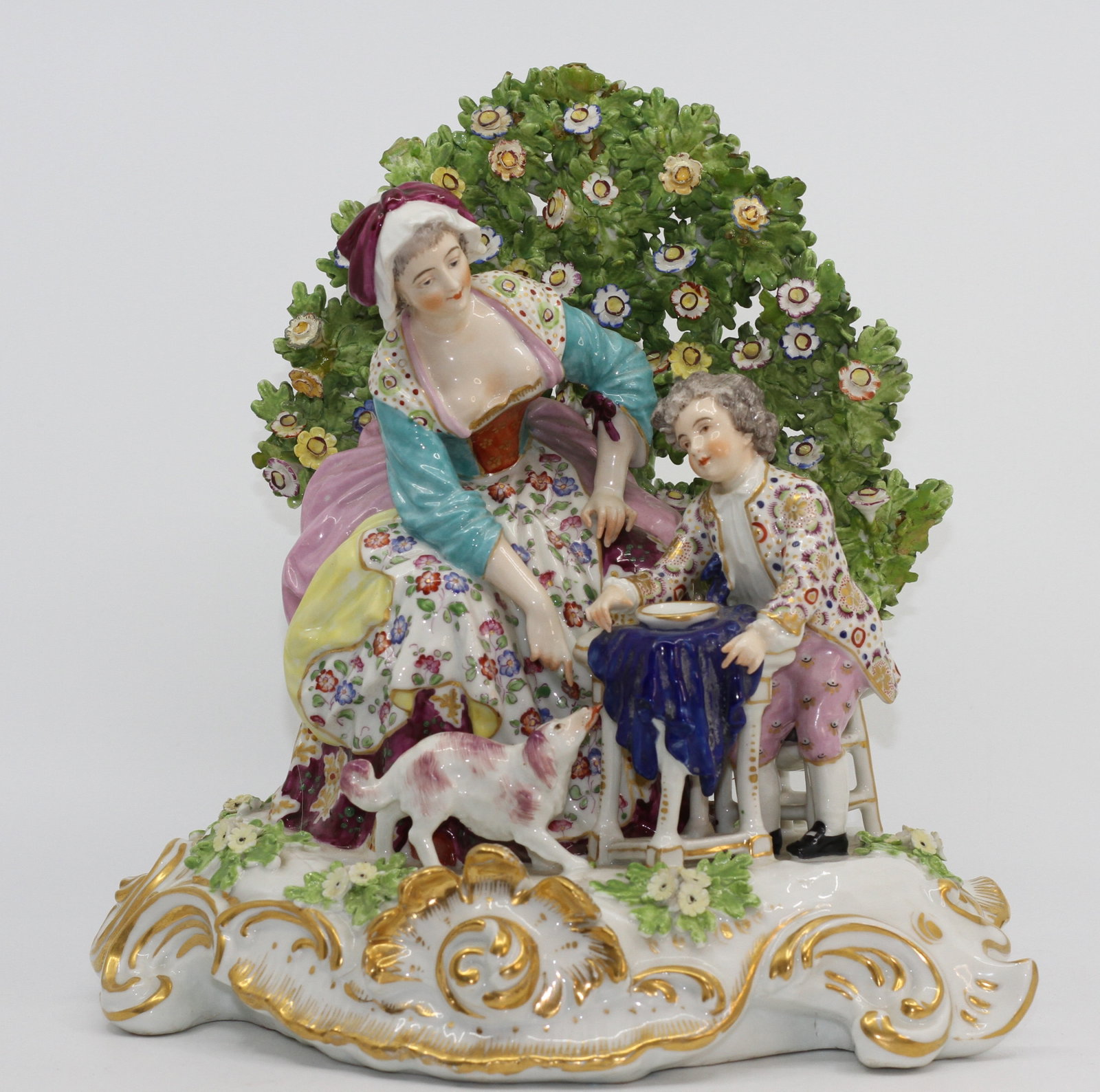 Antique Continental Porcelain Figural Group with Couple and Dog: Antique porcelain figural group depicting an elegantly dressed couple seated at a small table with a dog at their feet. Elaborate floral bocage backdrop, colorful enamel decoration, and extensive gilt