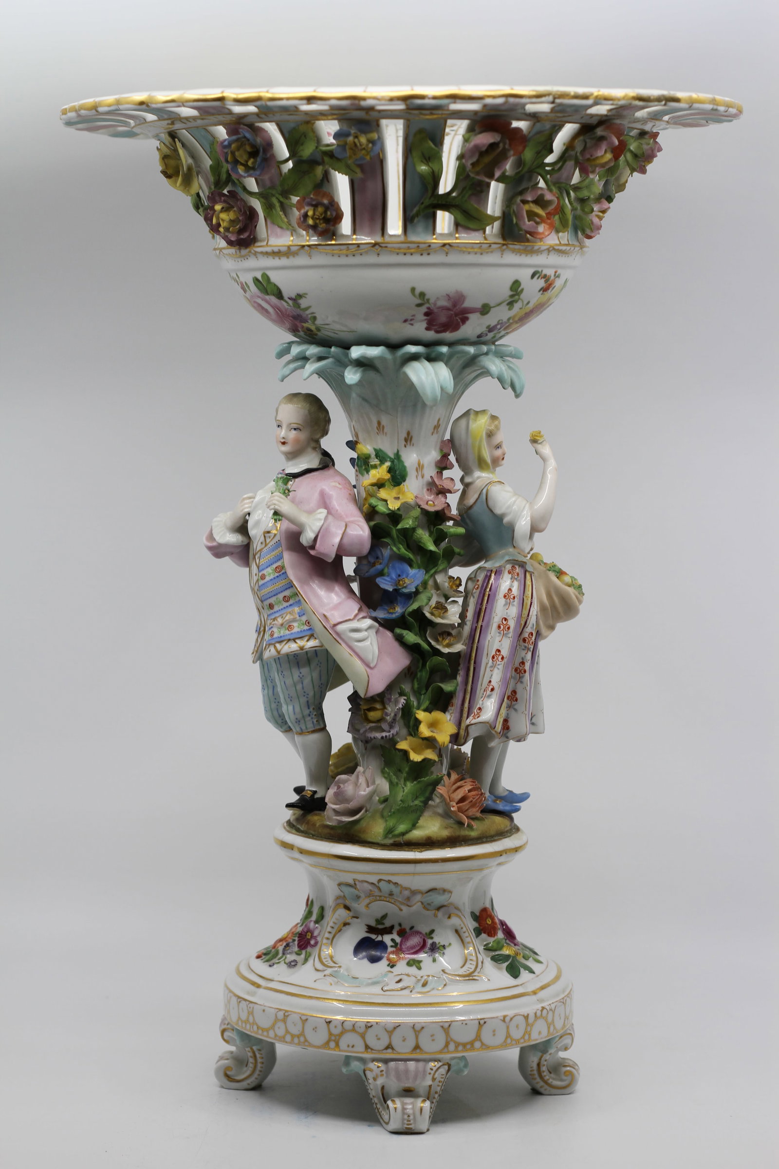 Antique Dresden Style Porcelain Figural Centerpiece: Large elaborate porcelain centerpiece or compote featuring a courting couple standing among applied flowers beneath a pierced floral basket. Hand painted with colorful floral decoration, gilt accents,