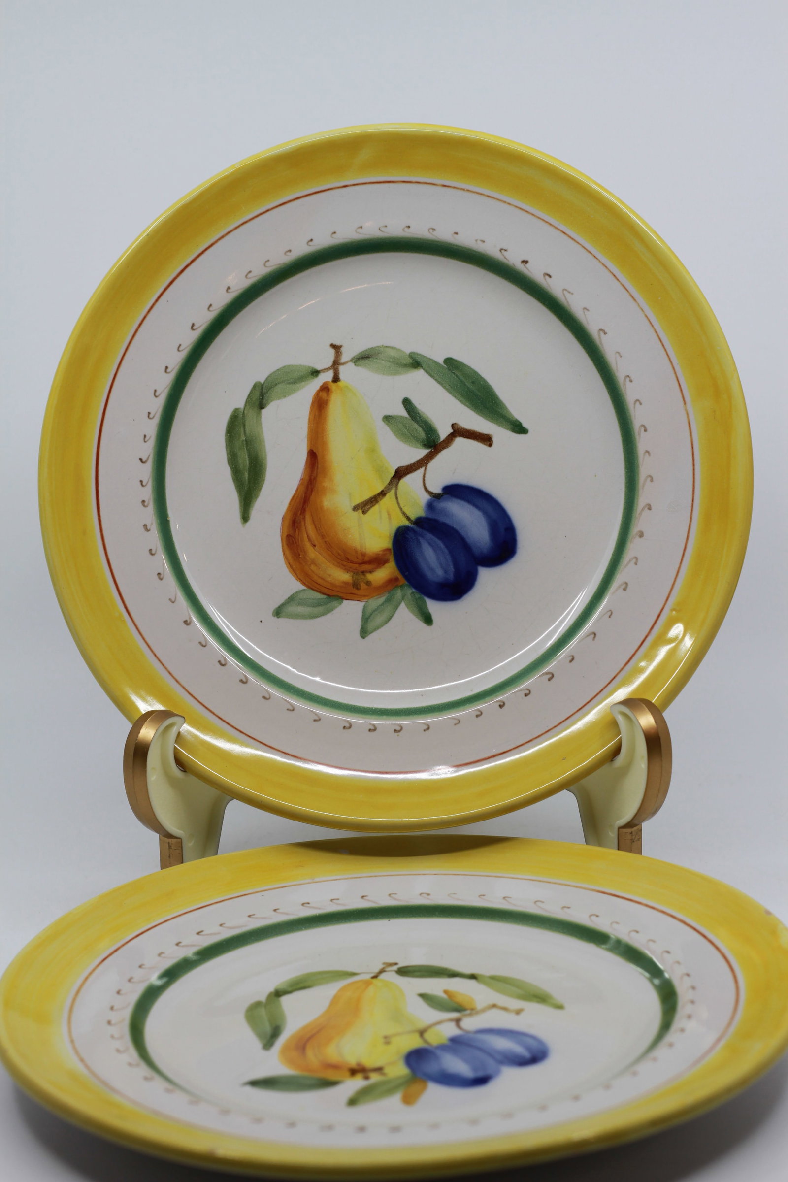 Pair of Hand Painted Fruit Decorated Porcelain Plates: Pair of porcelain plates decorated with hand painted pear and plum motifs, accented by green circular borders and bright yellow rims. Each bears a blue underglaze monogram style factory mark to the re