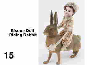 Bisque Doll Riding Rabbit: Germany, bisque head doll with brown glass eyes, open mouth with 4 teeth, incised 1902 12/0? on back of head on wood body with composition feet dressed in satin and tulle clown suit atop a flocked