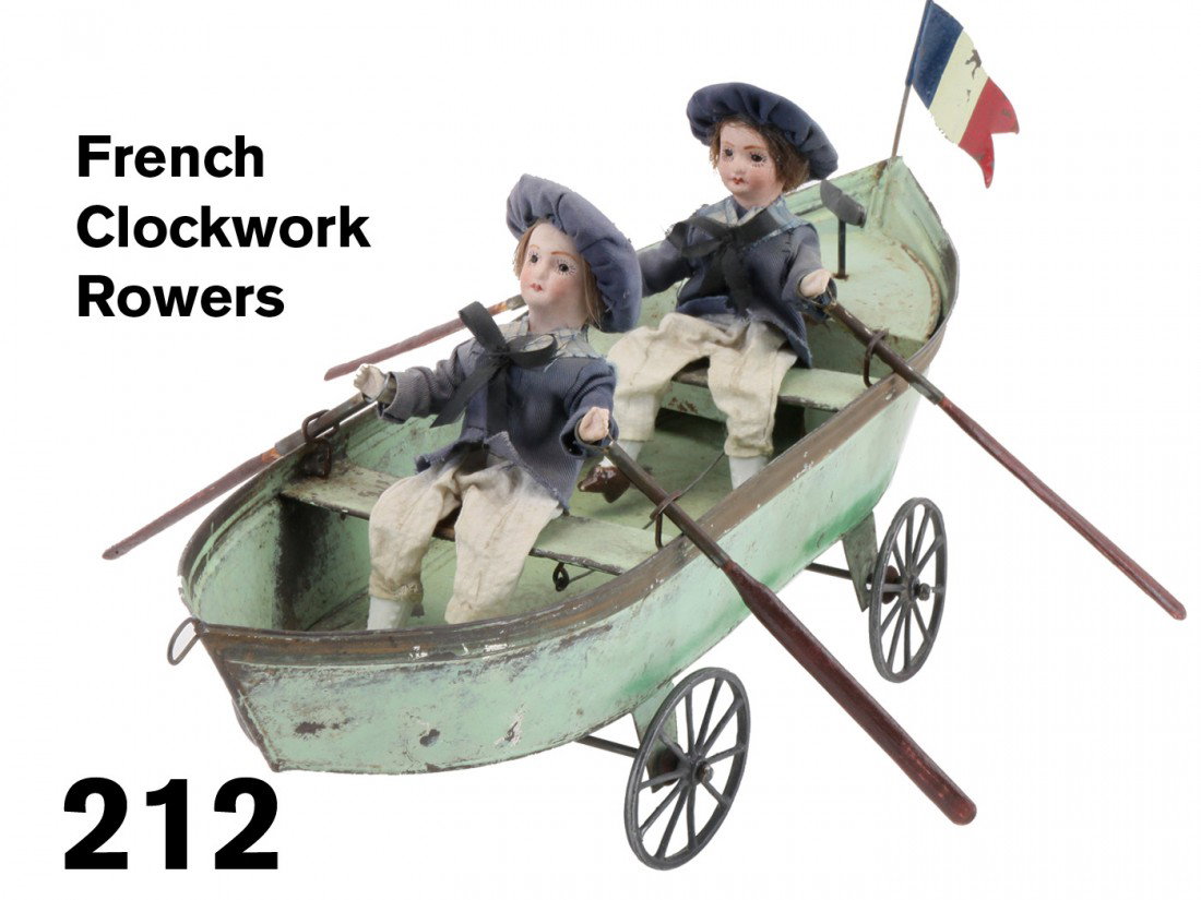 French Clockwork Rowers: two cloth dressed boy dolls with bisque heads and hands and china feet ride in a painted tin row boat with cast metal wheels, the stern compartment is adorned with a French tricolor banner and houses 