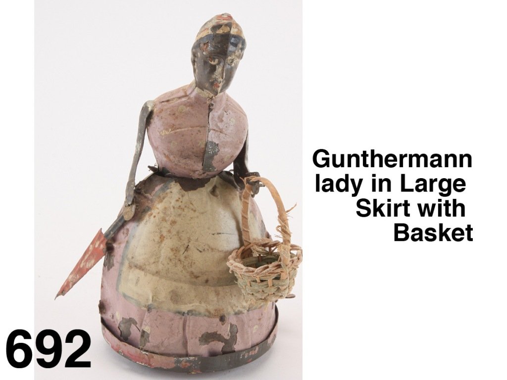 Gunthermann lady in Large Skirt with Basket: painted tin figure with internal clockwork, a walking figure who carries a basket and a furled polka dot umbrella Size: 6.5" t.