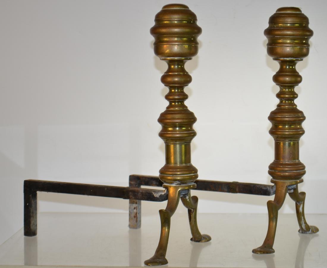 Pair of Brass Andirons: Andirons measure approx. 13"h. They have a solid brass body with cast iron posts and show common wear consistent with age.
