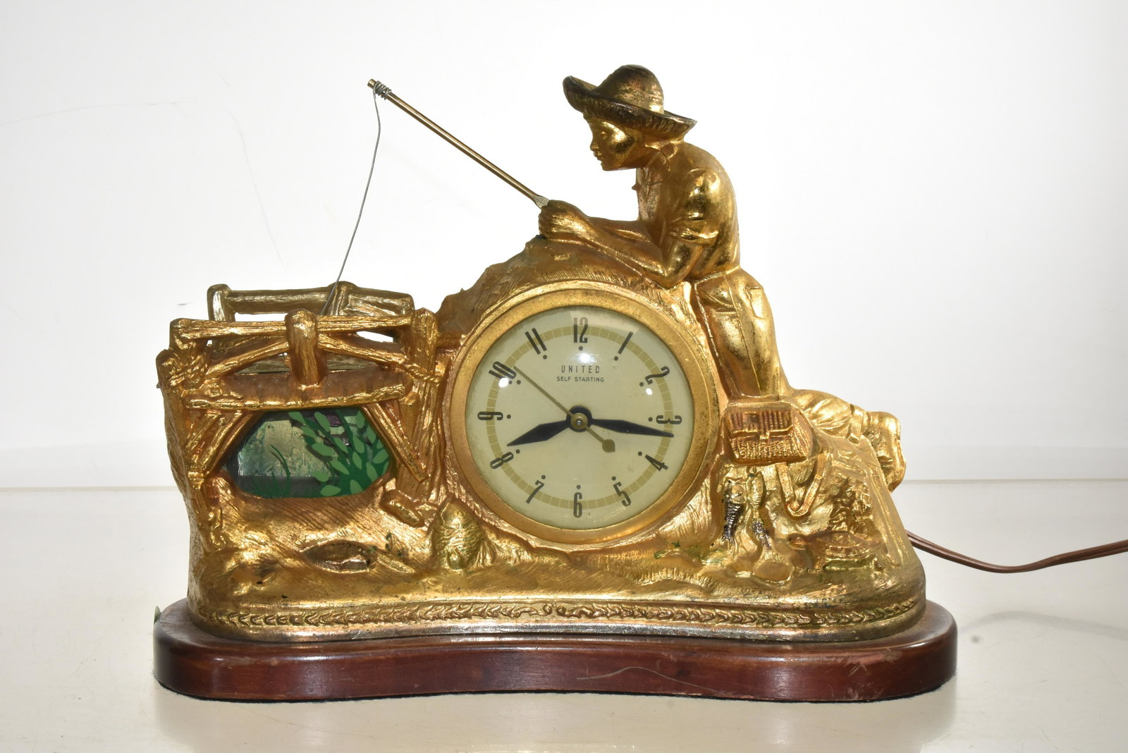 United Working Mechanical Fisherman Clock: Measures approx. 9 1/2h x 12" in length. Item is a United self staring electric clock. It depicts a young man fishing at a bridge. Clock has a glass window with painted water plants and behind the gla