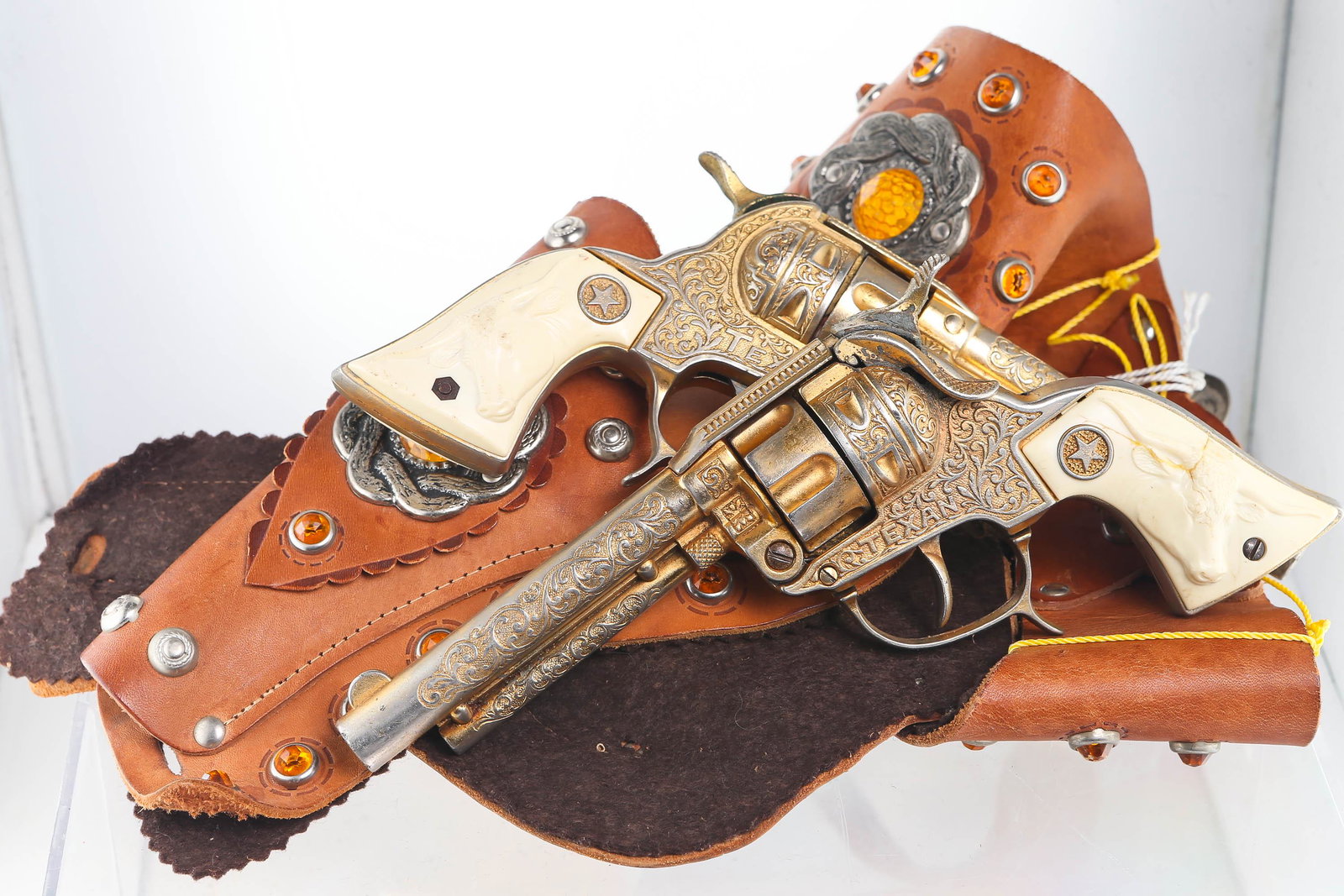 Texan Revolver Cap Guns, Pair in Double Holster: Texan revolvers have a gold tone with some wear, good action, barrel latch tight, handle has bas relief of steer on white plastic. Holster is leather with felt backing featuring many faux amber accent