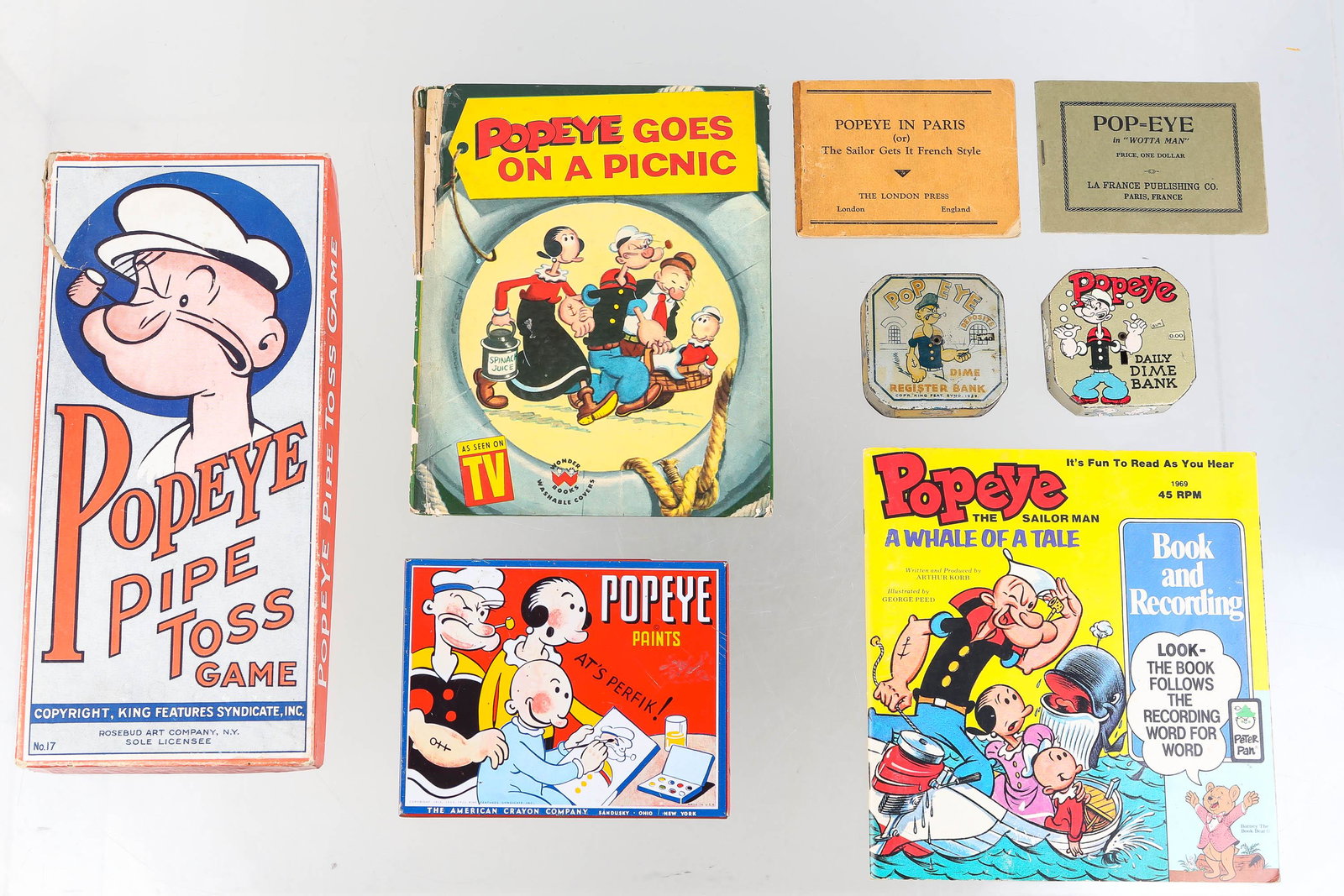 Vintage Popeye Banks and Collectibles: Lot includes 2 Popeye dime register banks, Popeye pipe toss game with Popeye figure, pipe and three rings (catalog No 17), Popeye watercolors, Popeye record (1969), 2 miniature Popeye comic books (one