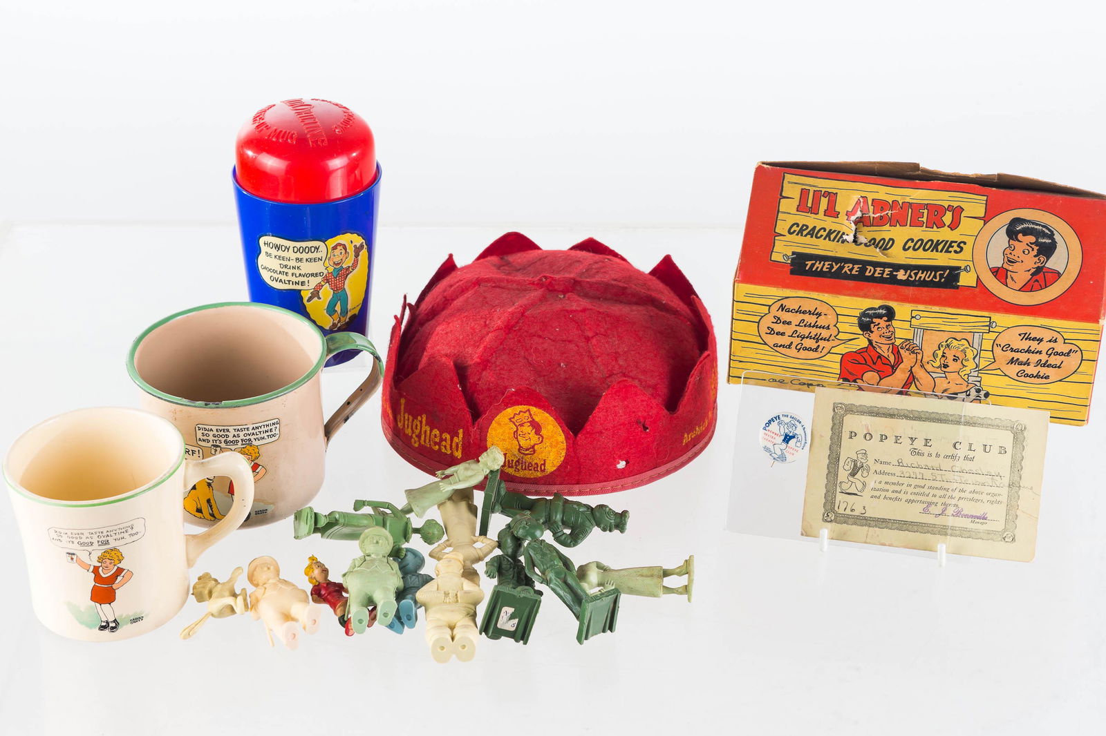 Mixed Cartoon Character Grouping: Lot includes a Lil' Abner cookie box with puncture, a Popeye Club pin back with club card, Ovaltine Howdy Doody shake up mug, Archie's Club jug head cap (1962), two Orphan Annie mugs (one is enamelwar