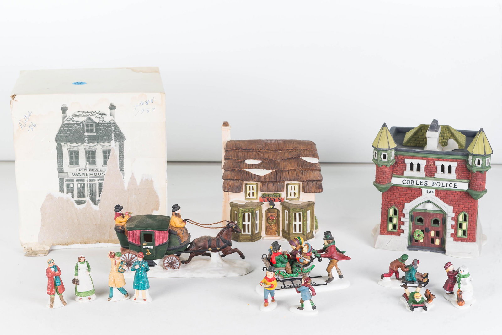 Studio 56 Dickens Village Collection: Lot includes 7 pieces, including Cobles Police Station, horse with coach, Caroling with the Cratchit Family, set of three snow children, set of 4 people, Fezziwig's Warehouse, and set of three shops.