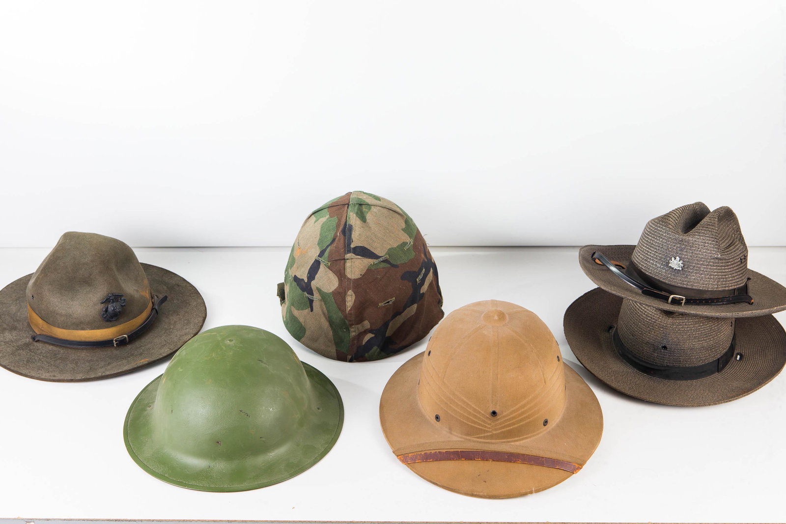 Military Helmets and Hats: Lot includes doughboy helmet, Korea War helmet, Civil Defense cloth helmet, Marine Sgt. cap, and two State Police hats (one with emblem).