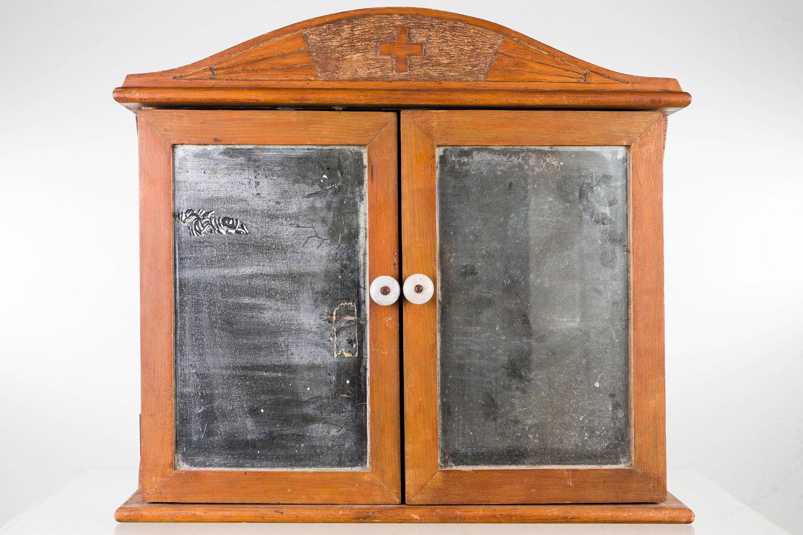 Vintage Two-Door First Aid Cabinet: Item measures approximately 18"H x 20"W x 6"D. Softwood cabinet with red cross symbol carved in crest. Item has two mirrored doors with wear, and multi section blue interior with a single drawer.