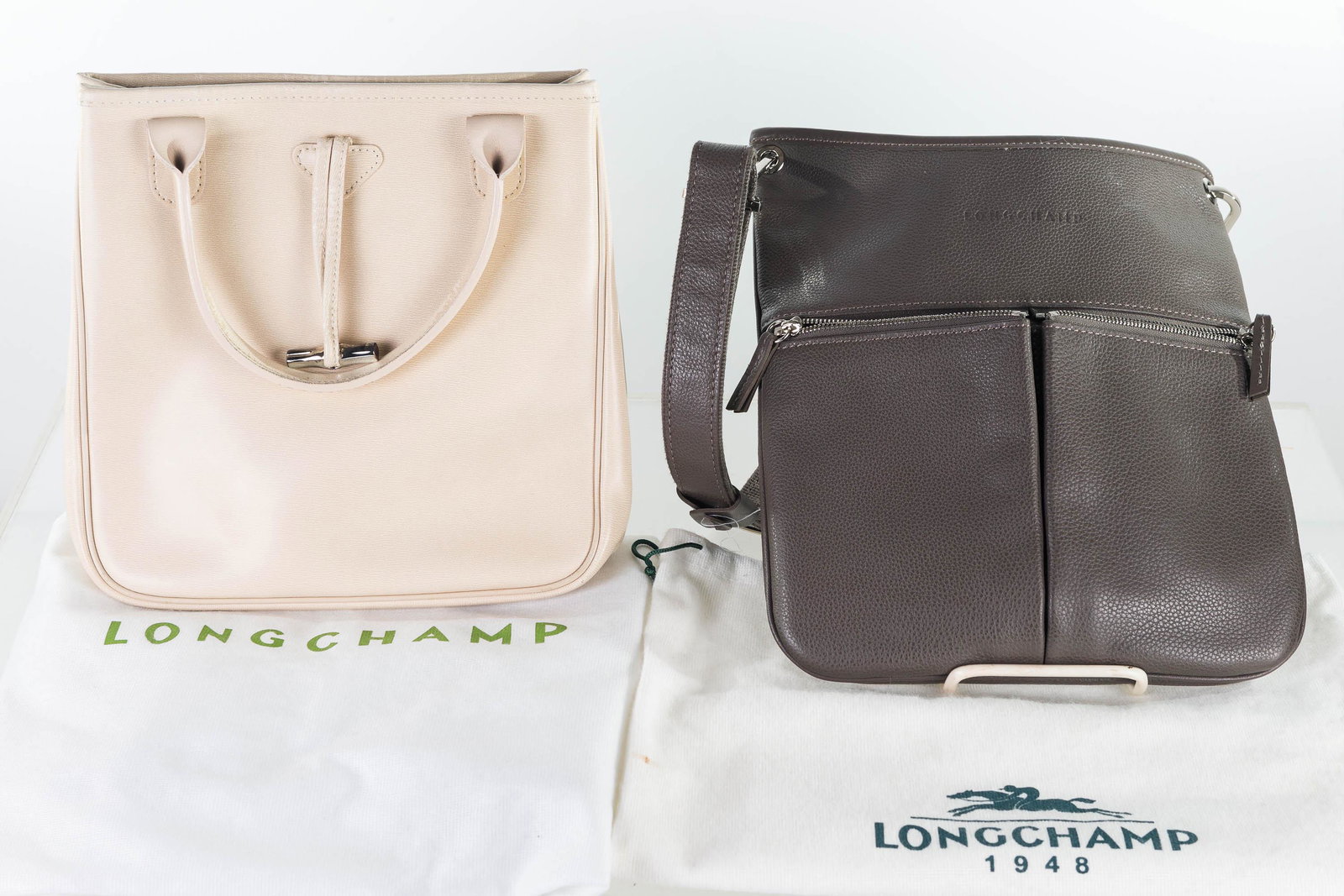 Longchamp Leather Purses: Both approximately 10" across. First bag is light tan with Longchamp logo woven in interior and one very small interior stain. The second is a dark brown with two exterior zipper pouches, and logo imp