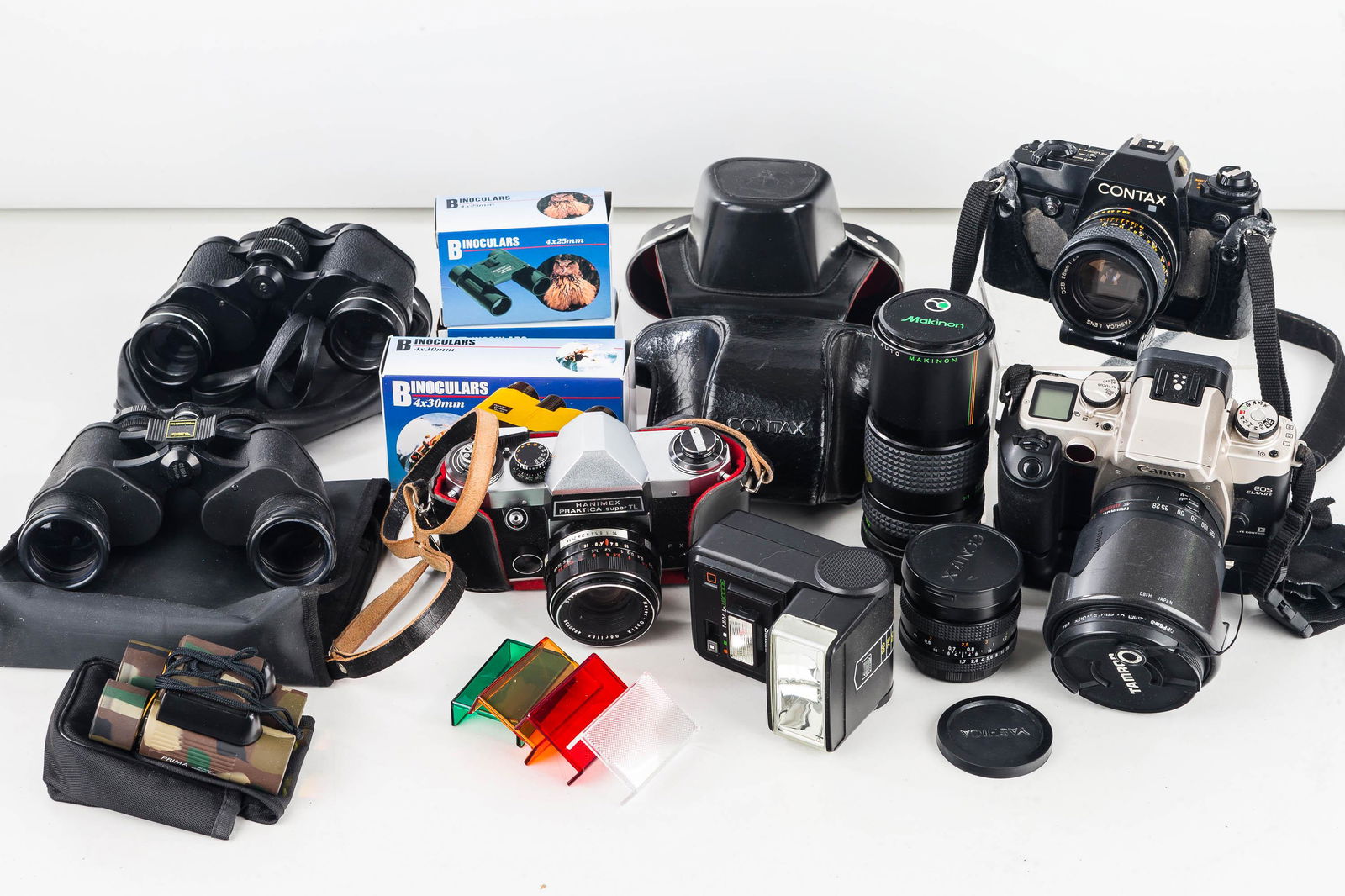 Vintage and Contemporary Cameras: Lot includes a Canon EOS with large Tamron lens, a Contax 35mm with roughness to body, a Hanimex Praktica Super TL 35mm camera, a small lens marked Carl Zeiss, a Makinon Zoom lens, and 6 pairs of bino