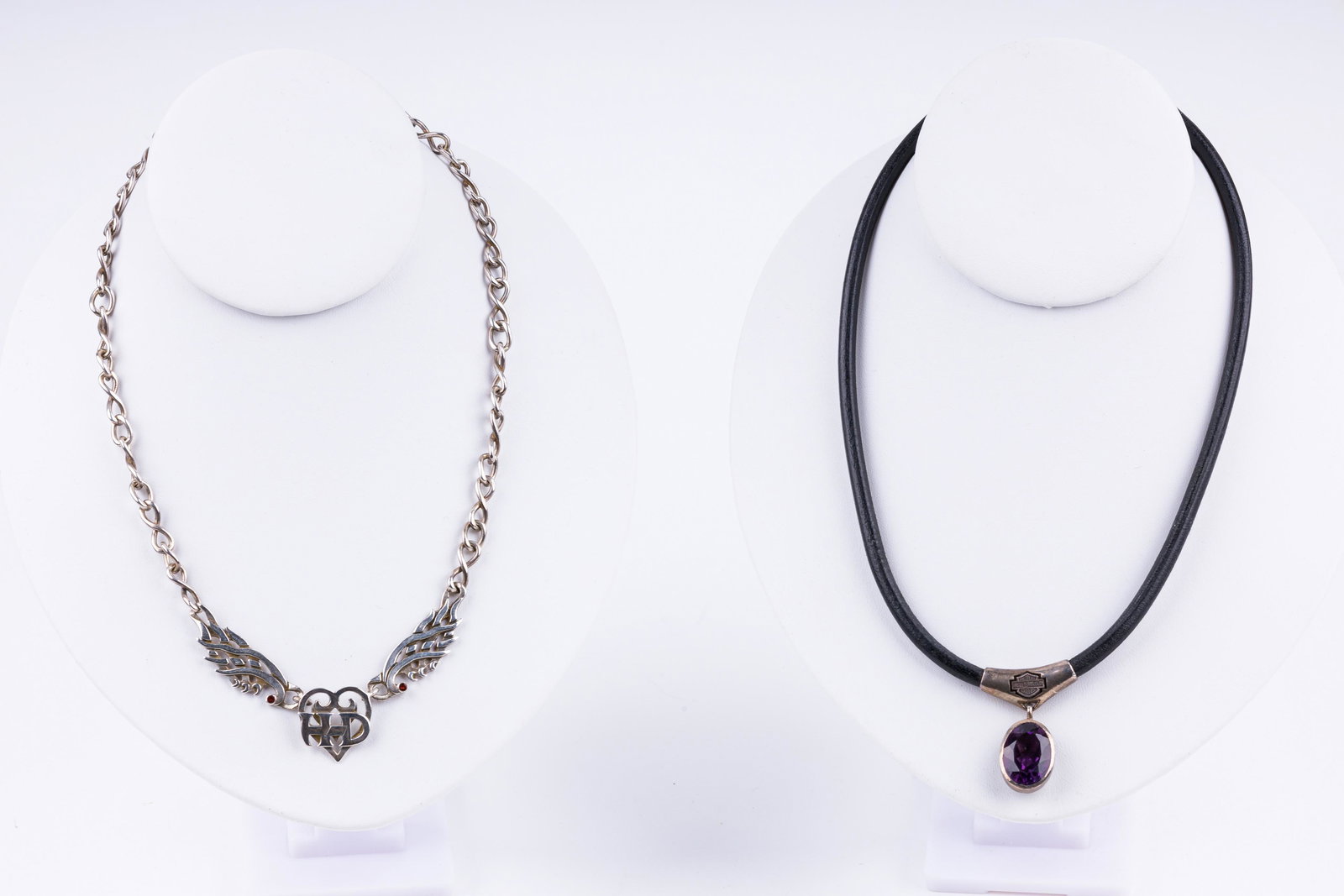 Harley Davidson Necklaces: Two Harley Davidson necklaces including one one leather cord and has central logo holding amethyst stone (approx 17.5" L) and chain with HD heart and wings with two garnets (approx 18" L).