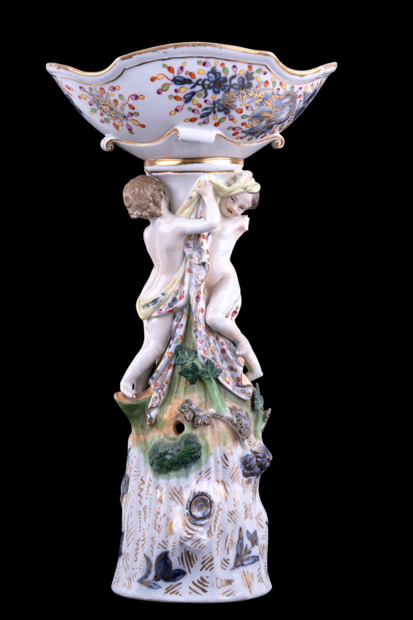 Meissen Porcelain Centerpiece (C. 1825-1924): A Meissen porcelain centerpiece measuring approx. 14.5" high with hand painted floral themes done in gold & multi-colored paint. Two robed females prop up bowl that measures approx. 8" across. One fem