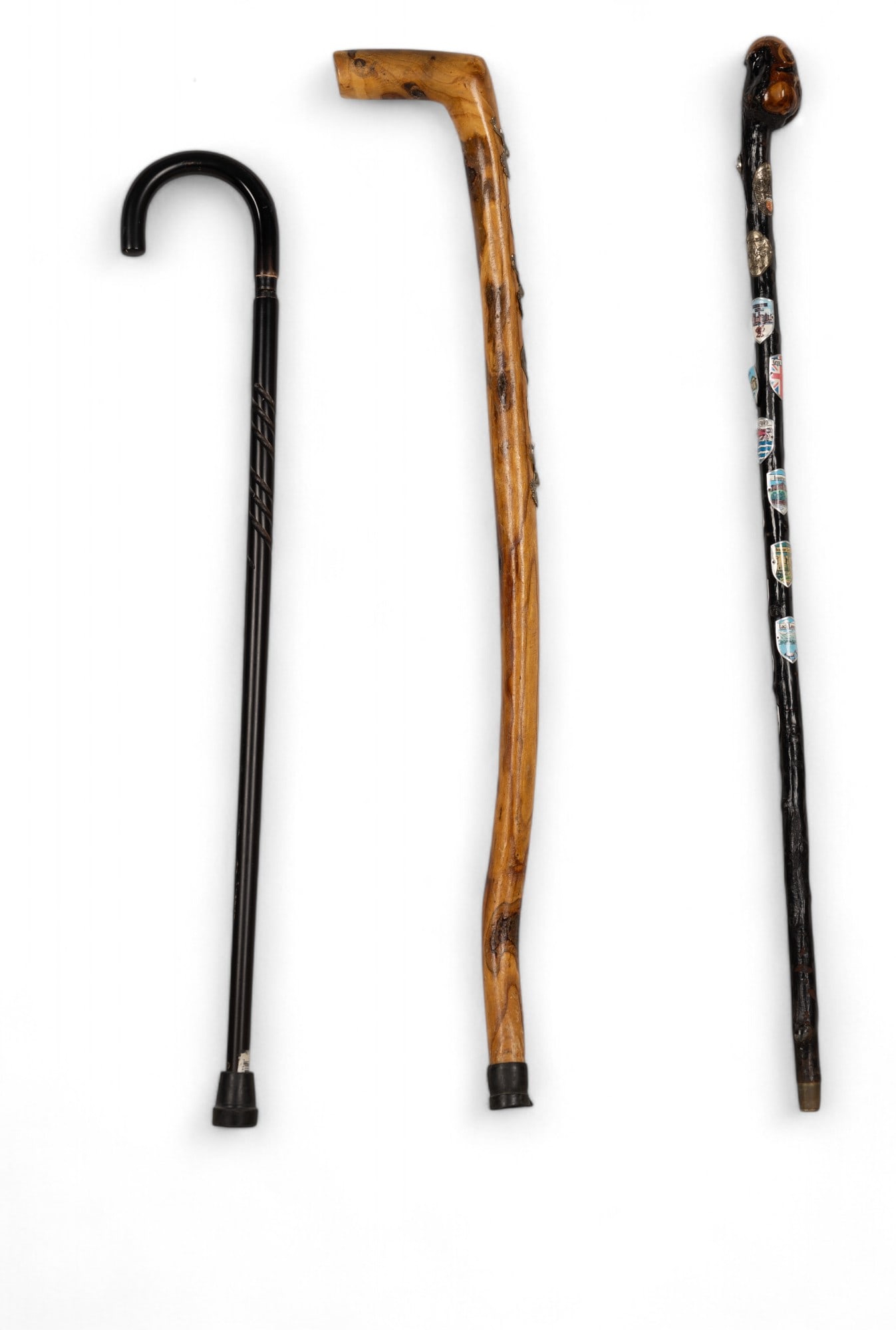 Travel Walking Sticks and Cane: 2 hand carved rustic walking sticks with assorted attached metal badges of destinations in the UK. 1 black wood cane with screw on curved wood handle and simple swirl carved design on shaft. All piece