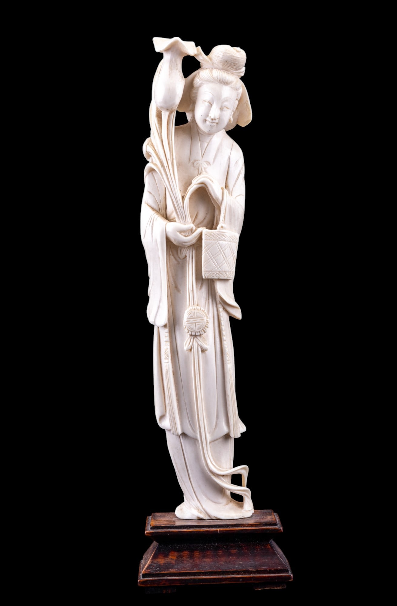 Chinese Guanyin Carved Bone Figurine on Stand: Chinese carved bone / natural material figurine of Guanyin, the bodhisattva of compassion, on a wooden stand. She is depicted holding a lotus bloom in her right hand & has a fish basket hanging on her