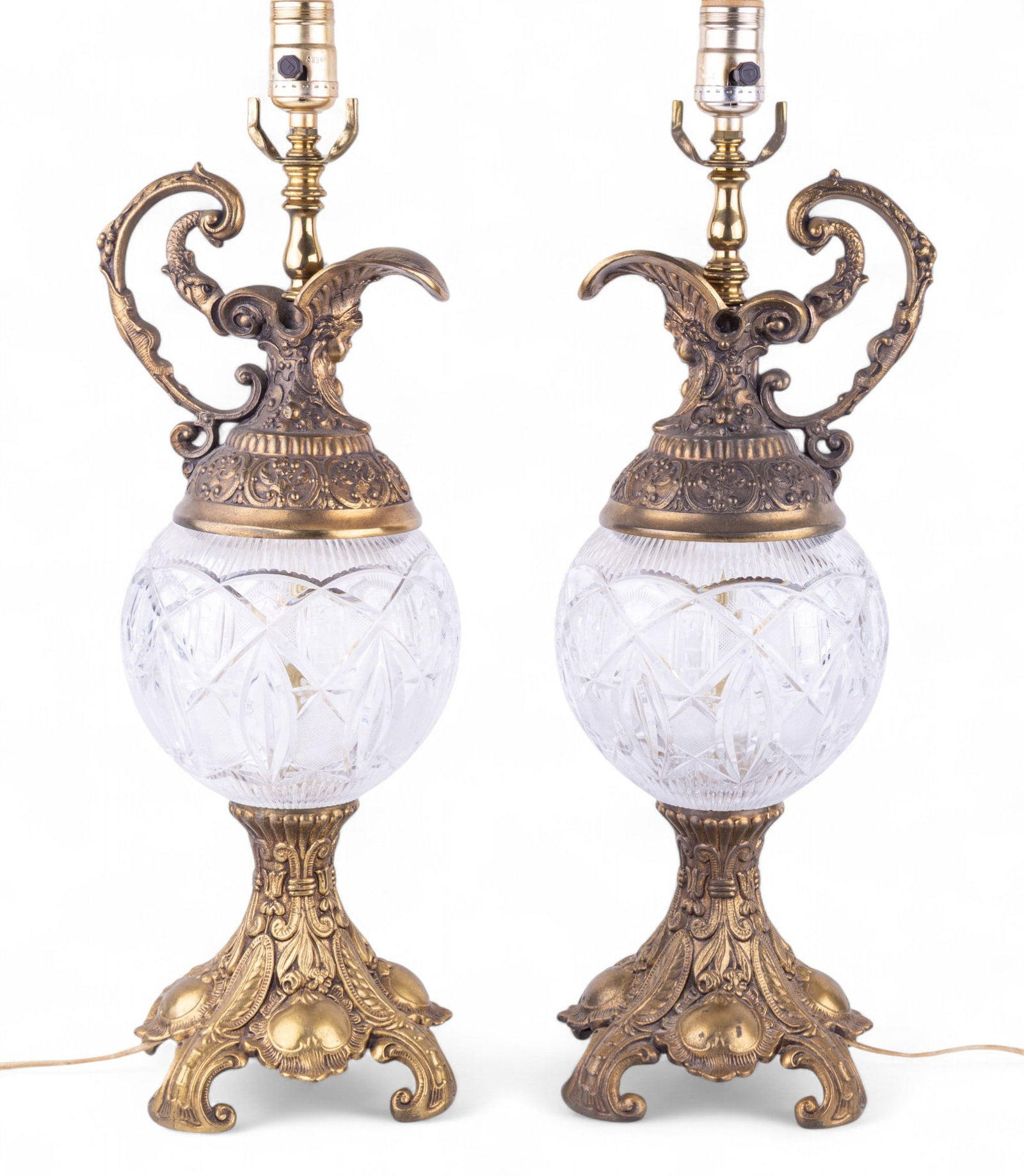 Pair of Early Rococo Style Crystal Globe Lamps: Pair of early Rococo style crystal globe lamps with higly decorated brass bases. Lamps are electrified. Cherubim faces, figural fish handle and more motifs. Measures approx. 33" h (with harp). Lamps i