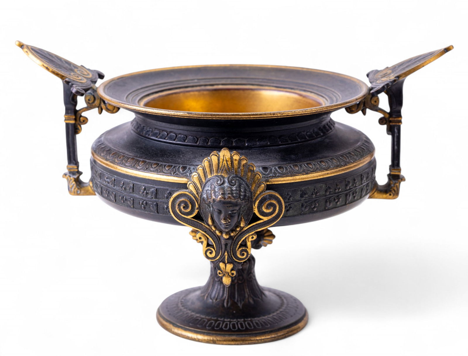 Bronze Egyptian Revival Tazza: Cast bronze tazza in the Egyptian Revival style, with dark patinated surface and gilt highlights. Features stylized female masks and ornate scrolled details on a circular pedestal base. Measures appro