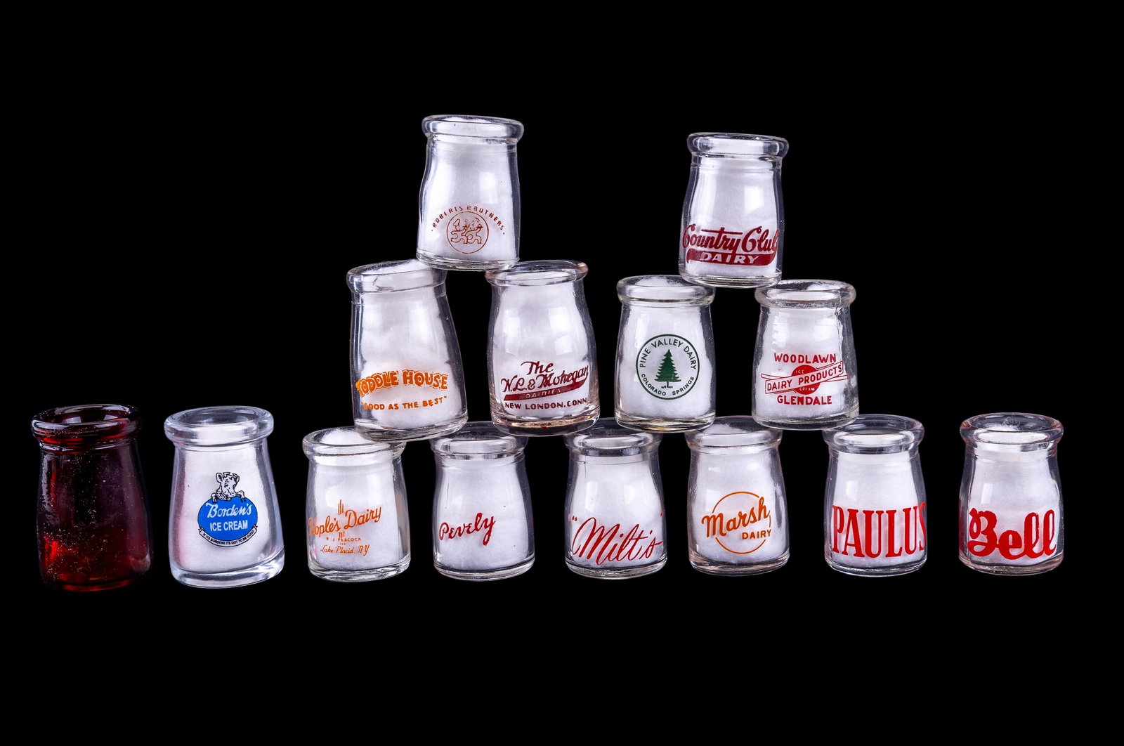 14 Vintage Dairy Creamers w/ Logos: Group of 14 vintage glass dairy creamers, each with original printed logos from various dairies and businesses including PAULUS, Bell, Pine Valley Dairy, and more. One red glass example.