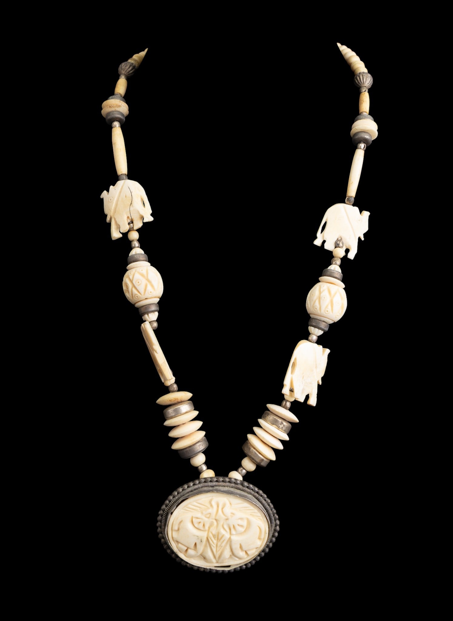 Carved Natural Material Elephant Necklace: An artisan carved natural material necklace featuring disc beads, round beads, carved beads, carved elephants, silver tone beads, and carved oval pendant, measures approx. 11" L on the drop.