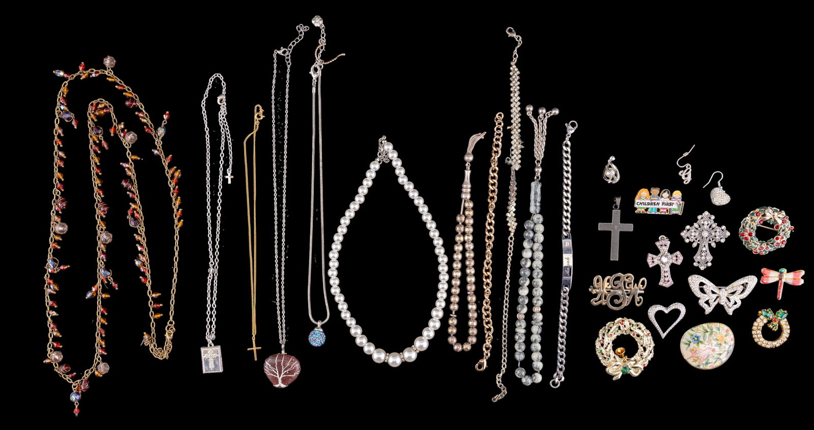 Fashion Jewelry Collection: Fashion jewelry displayed in a tray, including necklaces, bracelets, pendants, and brooches featuring various designs such as crosses, floral patterns, and beads. Materials likely include faux pearls,