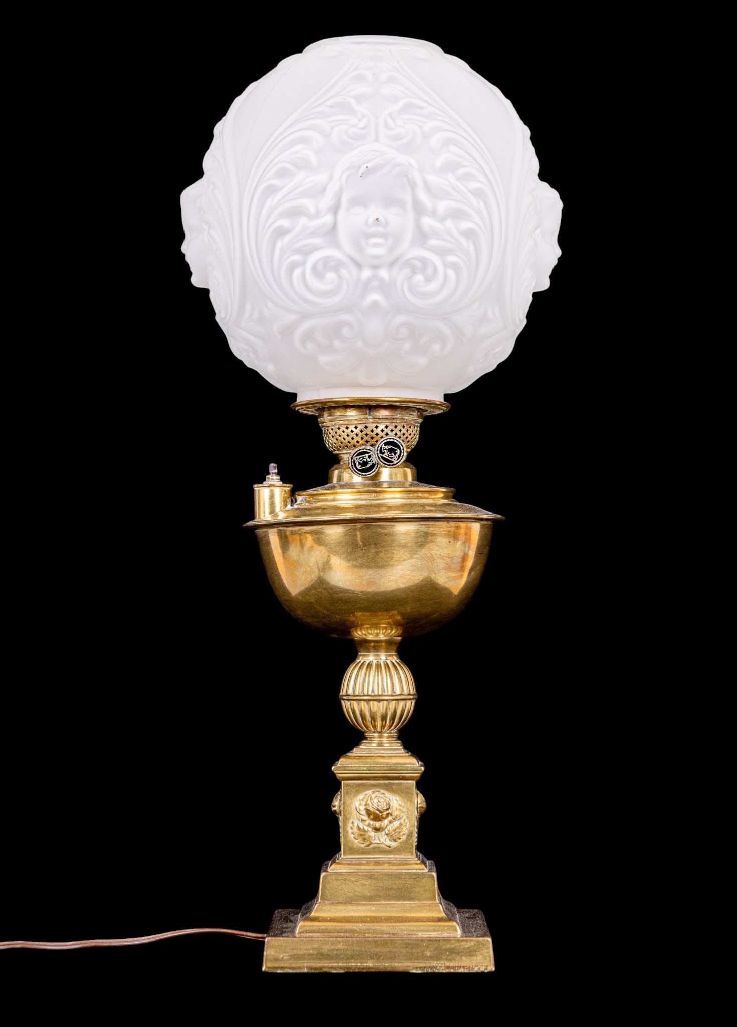 Early Brass Banquet Lamp: An early brass electrified banquet oil lamp conversion featuring rose relief details and a frosted glass cherub relief globe shade. Untested, note missing hurricane shade. Measures approx. 17" H to