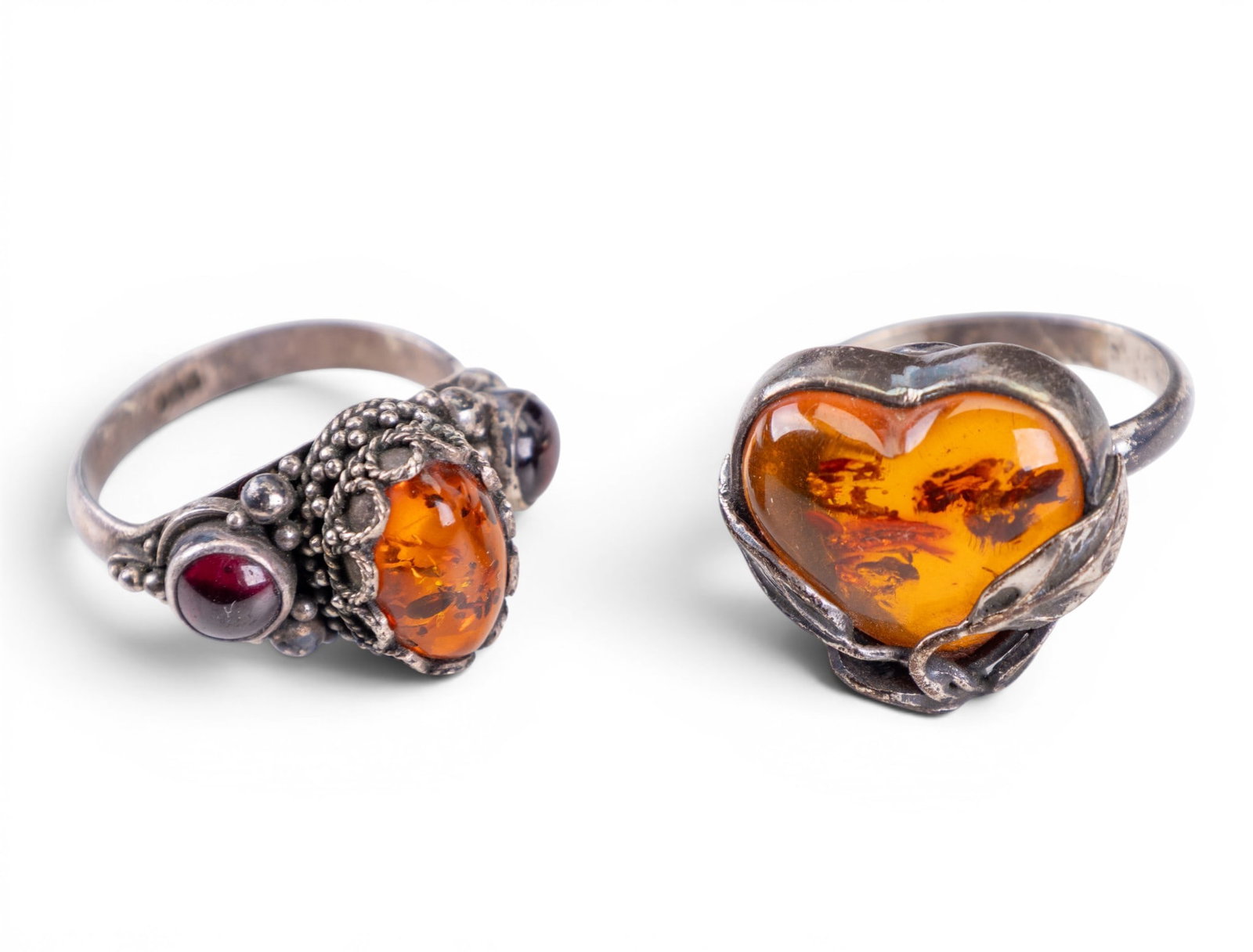 2 Vintage Amber & Sterling Silver Rings: A pairing of vintage amber and sterling silver rings including a figural heart amber sterling ring (size 8) and a housed amber/garnet sterling ring (size 9).