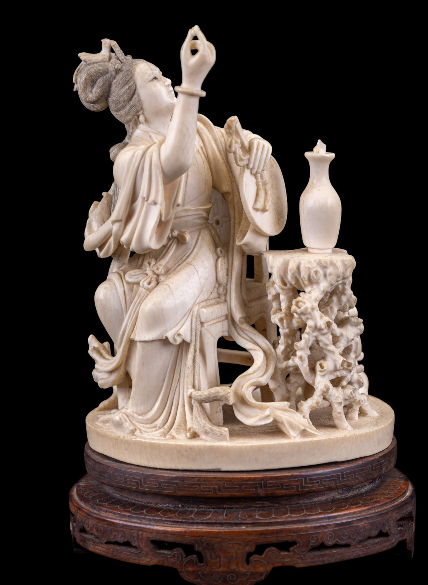 Chinese Hand Carved Figure of Seated Woman: Chinese carved natural material figure depicting a seated woman holding a musical instrument accompanied by a small table with vessel and additional decorative elements. The figure is rendered with in