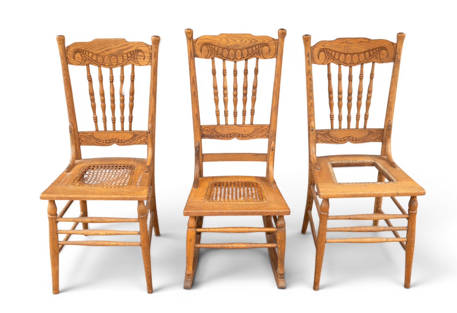 Cane Seat Spindle Back Chairs and Rocker: Two wooden spindle-back chairs and one rocking chair. Each features carved decorative motifs on the crest rail and a cane-woven seat, with visible wear and partial cane seat loss. Chairs measure 16