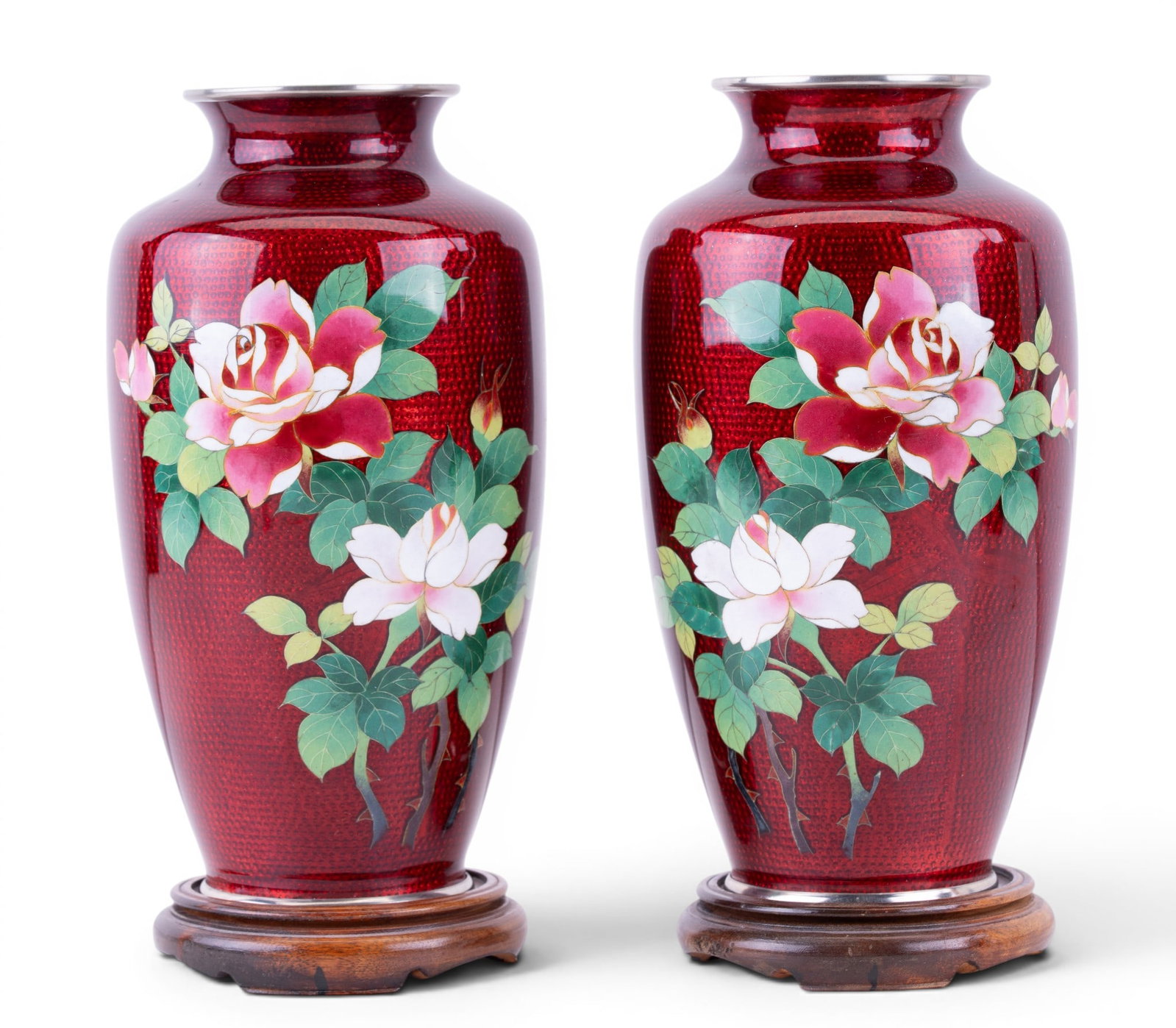 Pair of Chinese Enamel Floral Vases: Pair of Chinese enameled metal baluster-form vases featuring ruby red ground with hand decorated floral motifs of blooming roses and foliage. Each with silver-tone rim and foot along with a wooden dis