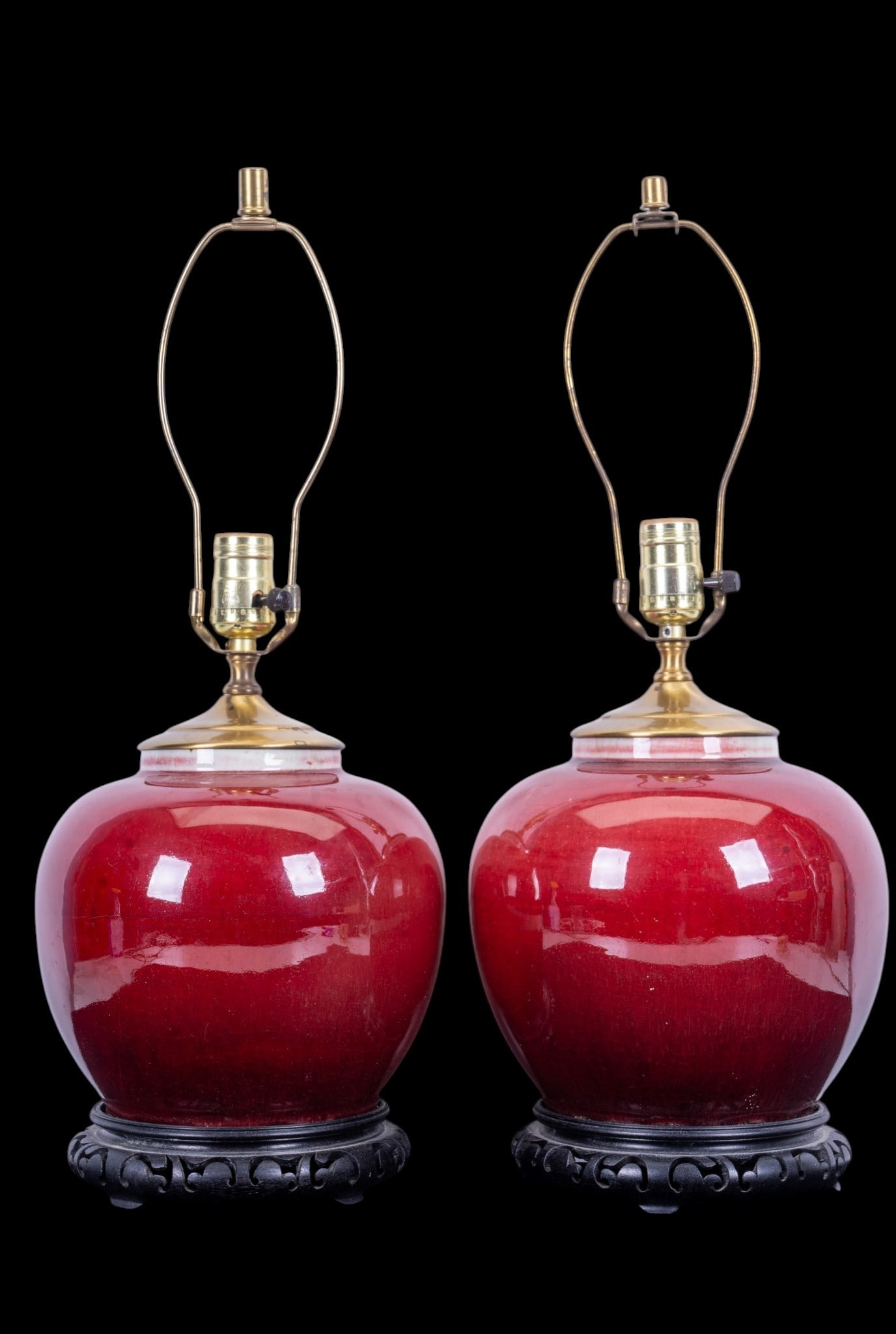 Pair of Chinese Oxblood Porcelain Vase Lamps: Pair of Chinese oxblood glazed porcelain vases converted to table lamps, each with a rounded body in red glaze. Mounted on carved dark wood stands. Each measures approx. 21" H to top of harp.
