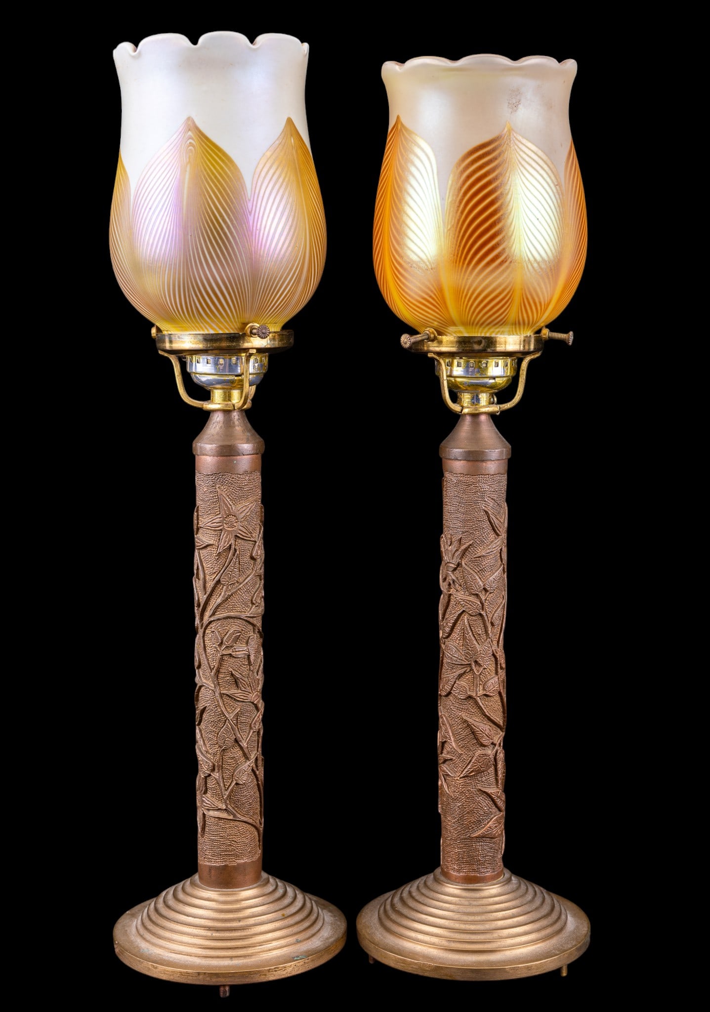 Quezal Favrille Shades and Art Nouveau Lamps: A pair of brass Art Nouveau electrified table lamps featuring a relief design of bird of paradise flowers up the pedestal. Included are two Favrille glass shades, one signed Quezal on the inner rim,