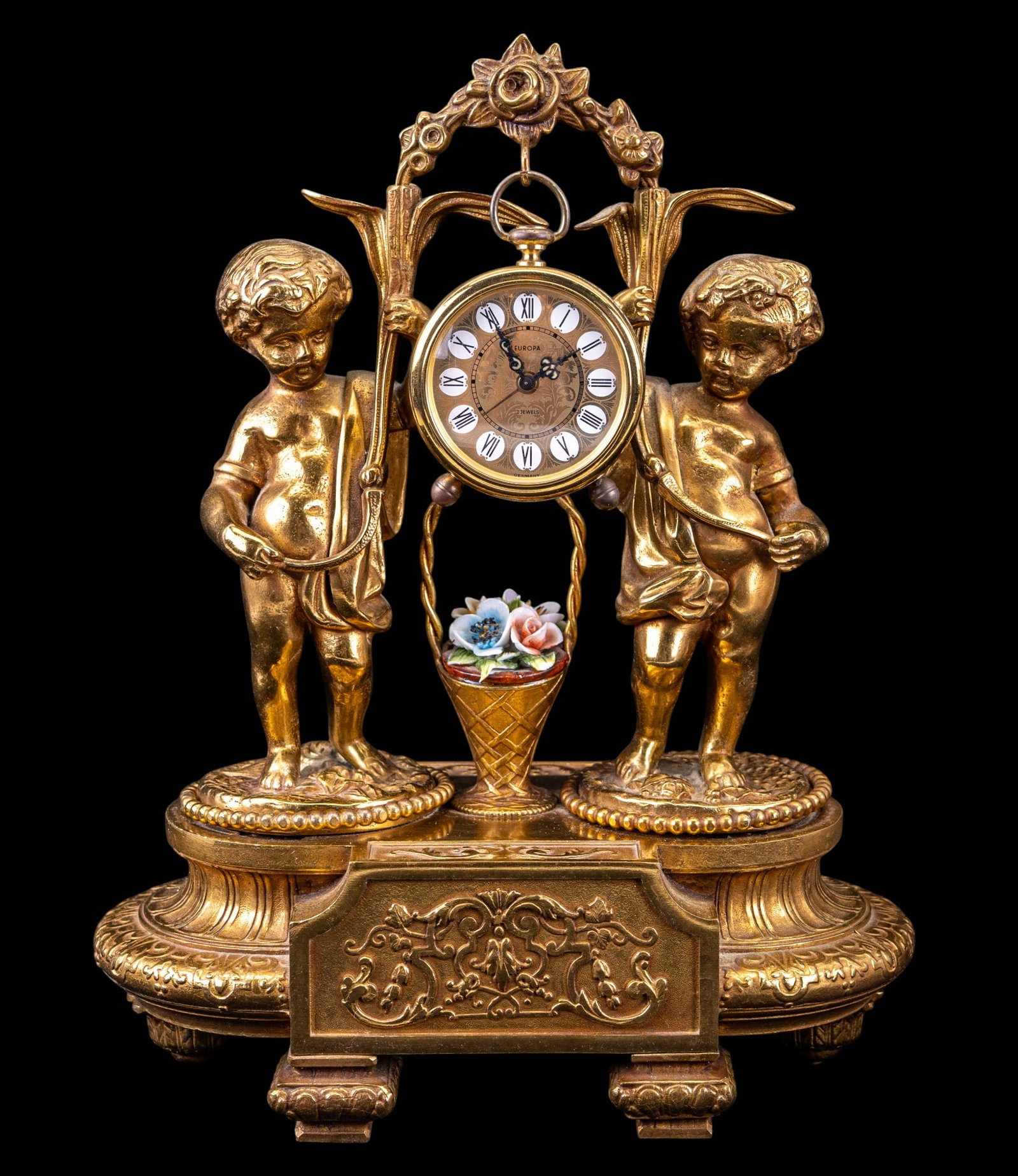 Italian Bronze Cherub Clock Stand & Europa Clock: An mid century figural bronze cherub desk top stand featuring cherubs and porcelain flowers, and a hook for hanging a small clock or pocket watch, marked 'F.B.A.I.' from the Fonderia Bronzi Artistici