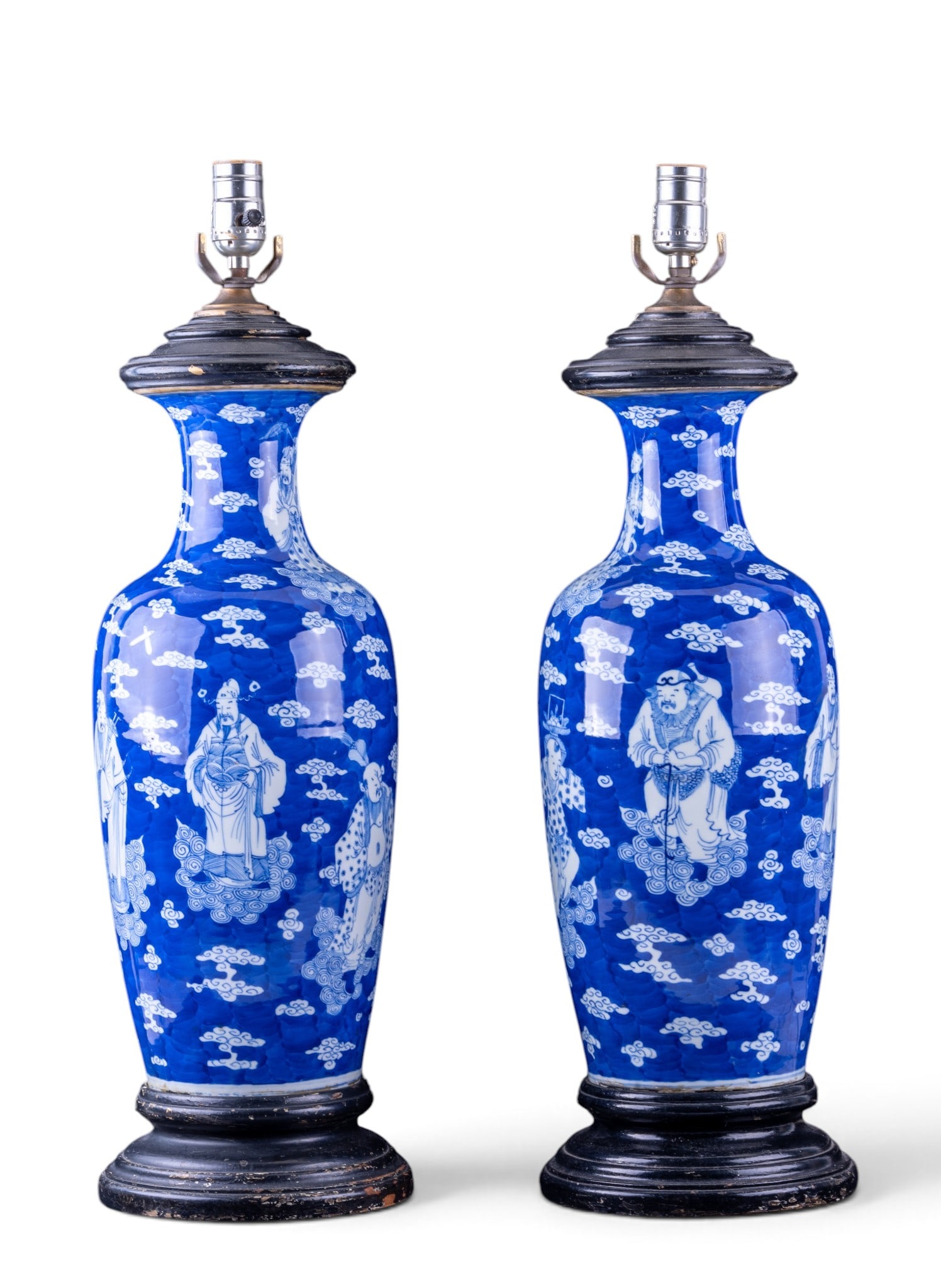 Pair of Chinese Blue & White Immortals Table Lamps: Pair of Chinese porcelain table lamps decorated in underglaze blue and white with immortals and figures among stylized clouds. Each mounted on a carved dark wood base with matching upper wood fittings