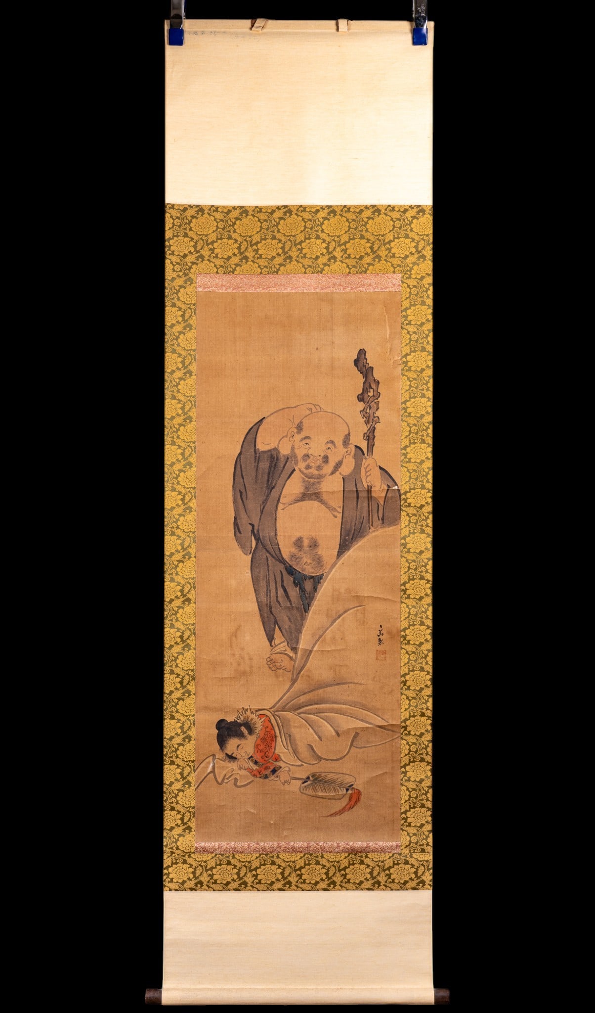 Japanese Hanging Scroll: Antique Japanese hanging scroll depicting Hotei (the Laughing Buddha) standing over an attendant or child sleeping on his massive sack of treasures. The right side features a stylized two-character ar