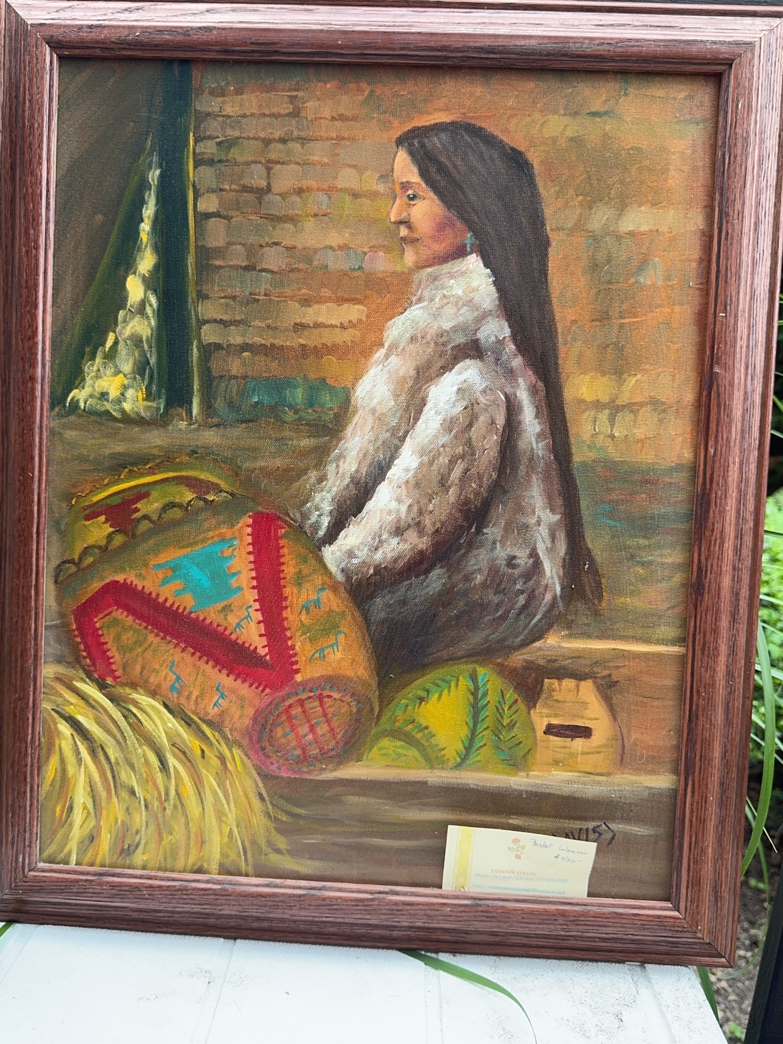 Connie Davis Oil Painting of Seated Woman: Original oil painting on canvas by Connie Davis depicting a seated woman with long dark hair, wrapped in a fur garment, beside vibrant patterned baskets. Signed lower right. Framed with a wood-effect 