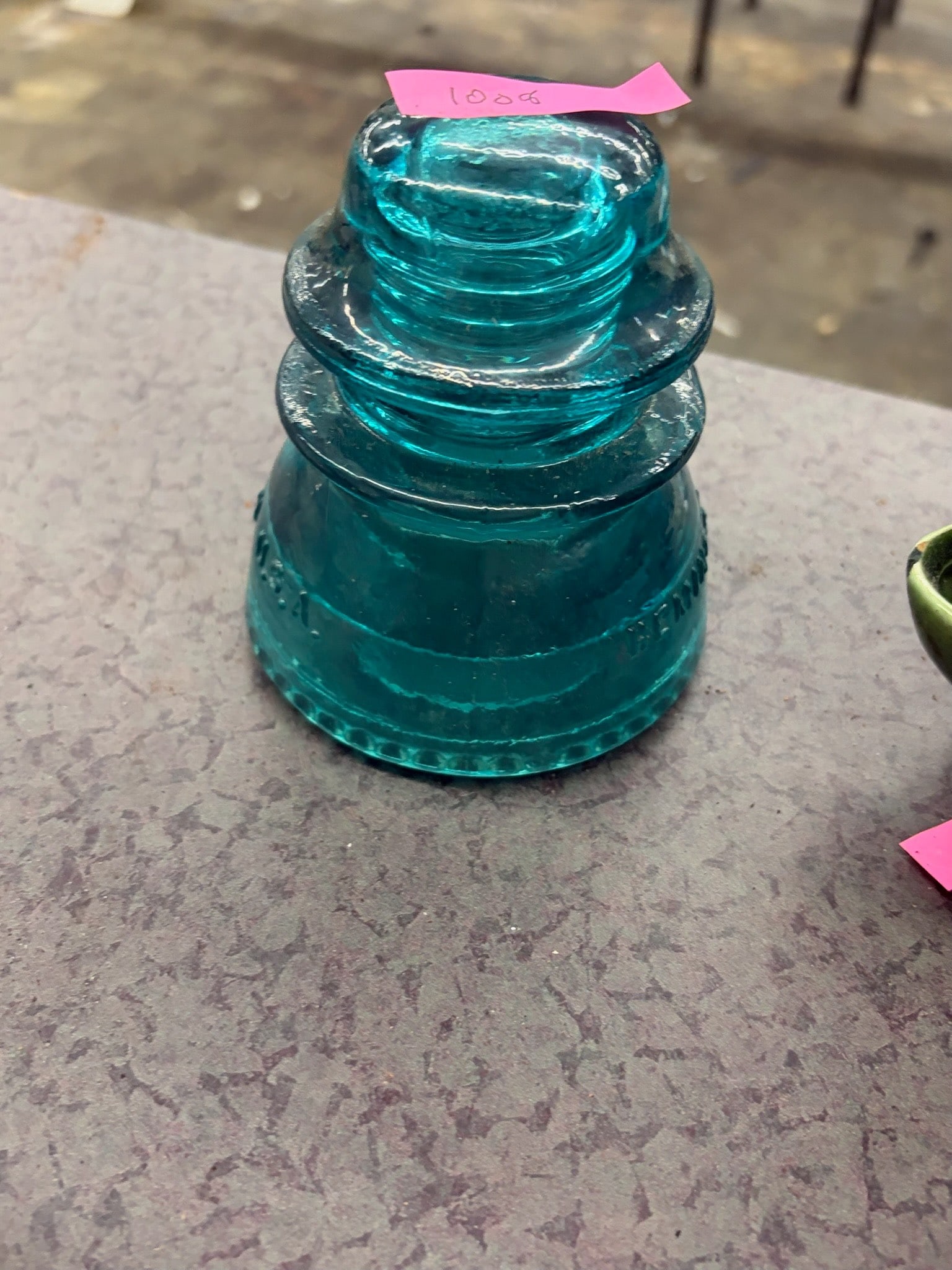Aqua Glass Insulator with Embossed Details: A vintage aqua glass insulator, featuring an embossed design. Commonly used in the mid-20th century for telegraph or electrical lines, it showcases a distinctive tiered shape and vibrant colored glass