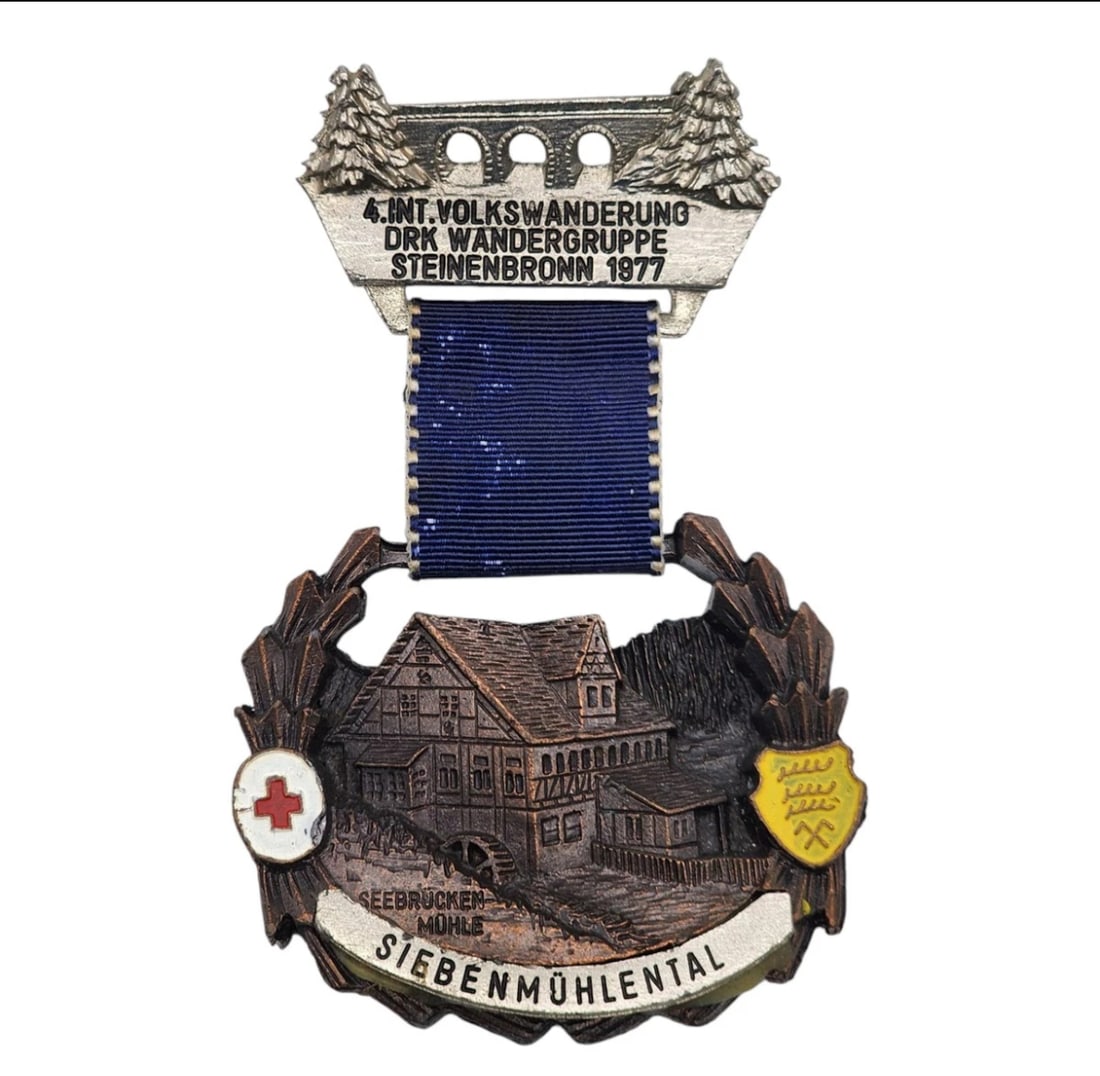 German Hiking Medal DRK Steinbronn Volkswanderung 1977 Siebenmohlental: highly collectible German commemorative volksmarch medal from 1977. The top silver-tone header bar features a relief design of a stone bridge flanked by evergreen trees, inscribed with "4. Int. Volksw