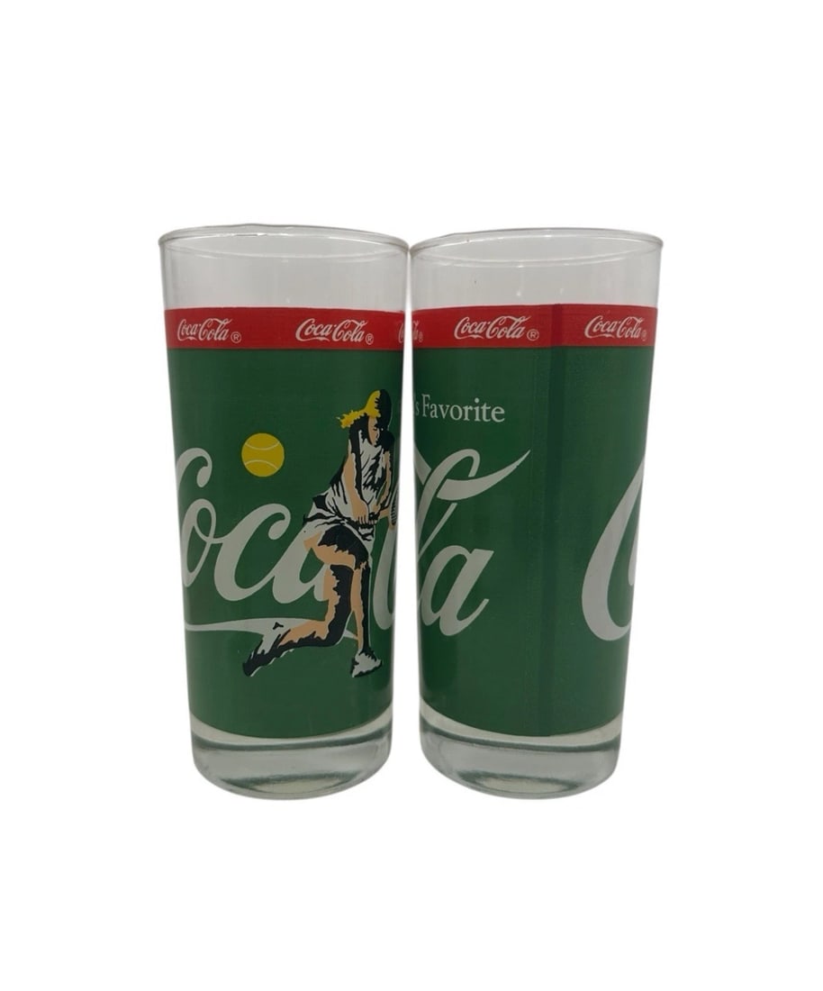 Vintage Set Of 2 Coca Cola Drinking Glass Female Tennis Player Glasses: A vibrant pair of vintage 1970s/1980s Coca-Cola promotional sports-themed tumblers. Designed with a solid forest green wrap-around background, these clear glass tumblers showcase a retro graphic of a 