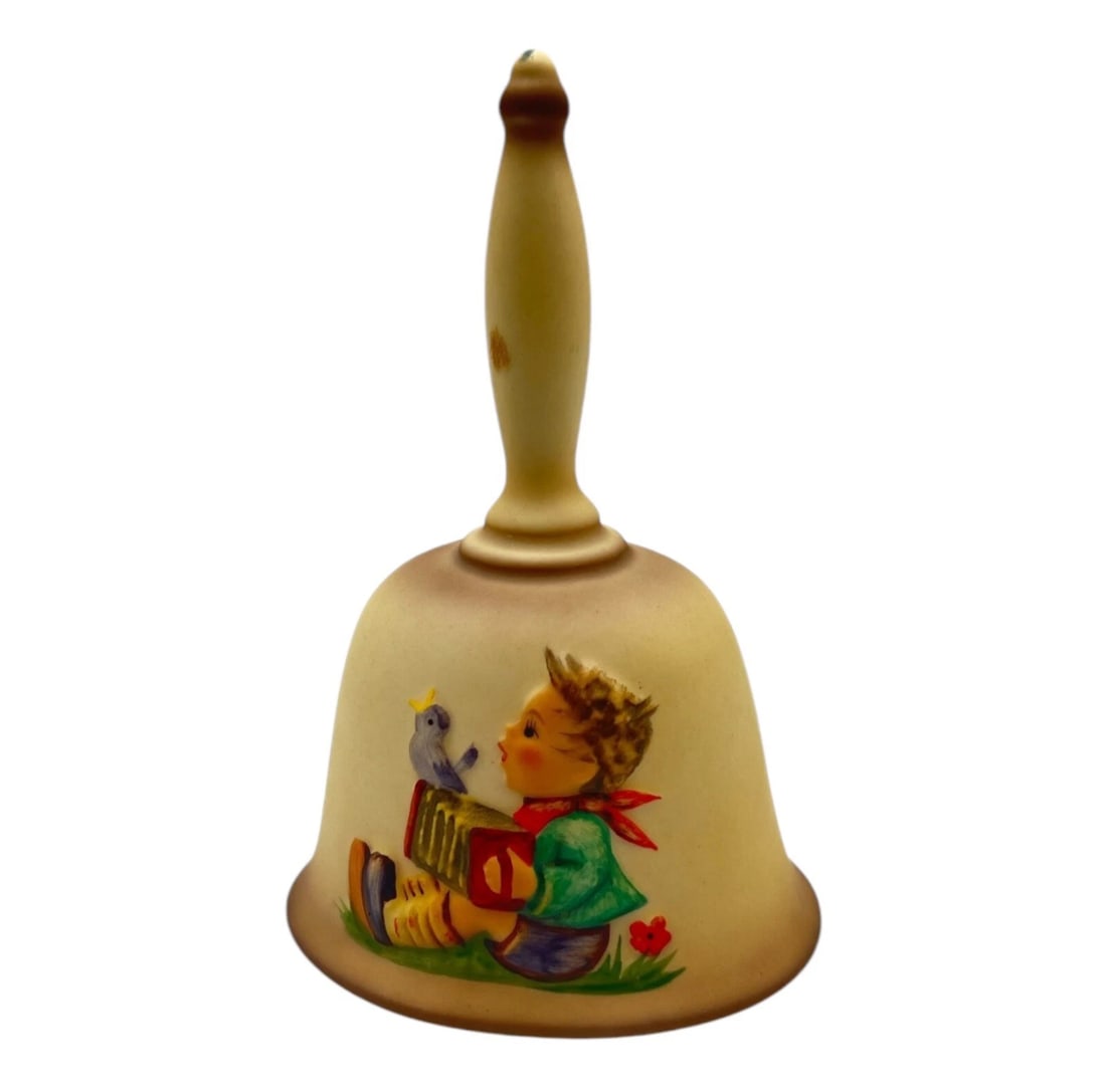 Vintage Goebel Hummel Figurine First Edition Annual Bell 1978 HUM700: ceramic hand bell featuring the iconic artwork of Sister Maria Innocentia Hummel. The face of the matte-finished bell displays a raised relief graphic of a young boy sitting in a field playing an acco