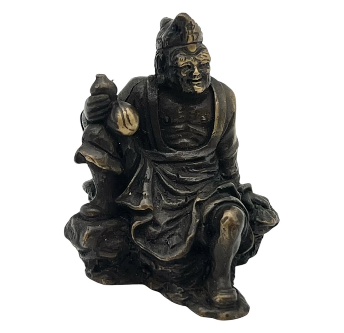 Vintage Brass Ji Gong Chinese Buddha Figurine: A small, patinated bronze figurine depicting a seated or reclining male figure with a highly expressive, wrinkled face, large ears, and a soft hat or head covering. He sits casually on an irregular, r